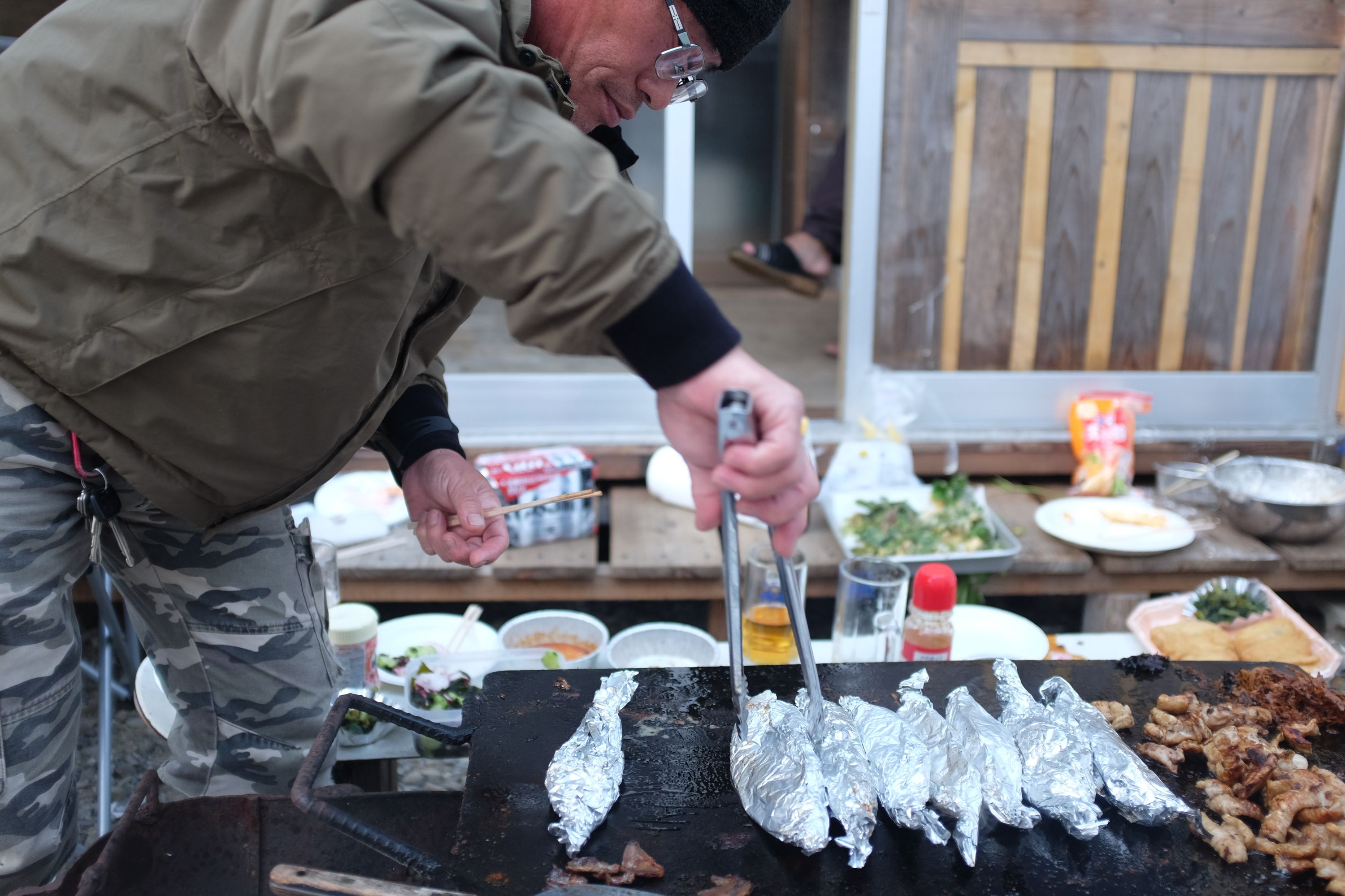 A man in a flight jacket and glasses turns small fish wrapped in aluminium foil on a grill.