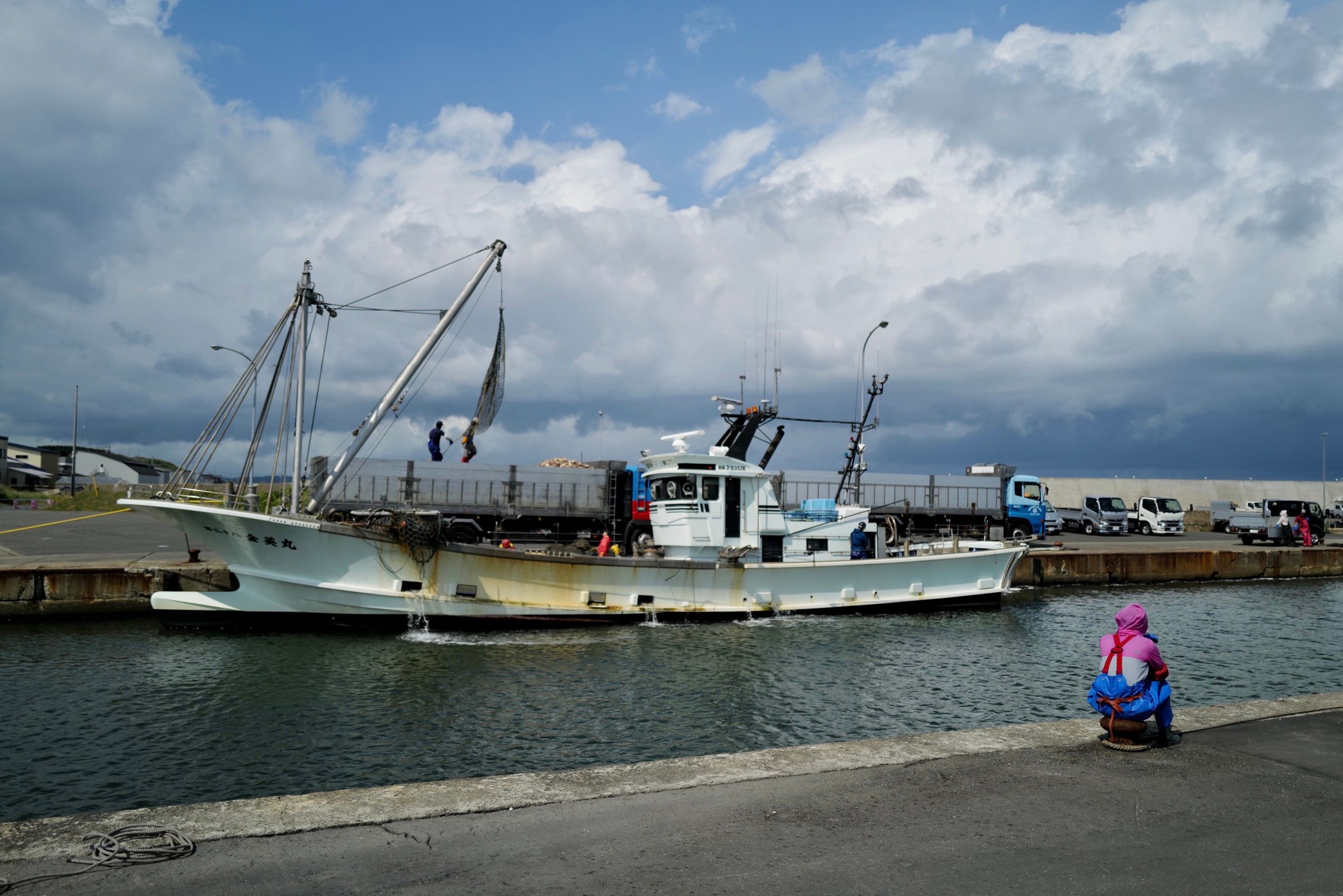 A woman in fishing overalls sits on a bollard a watches a scallop boat unload its catch