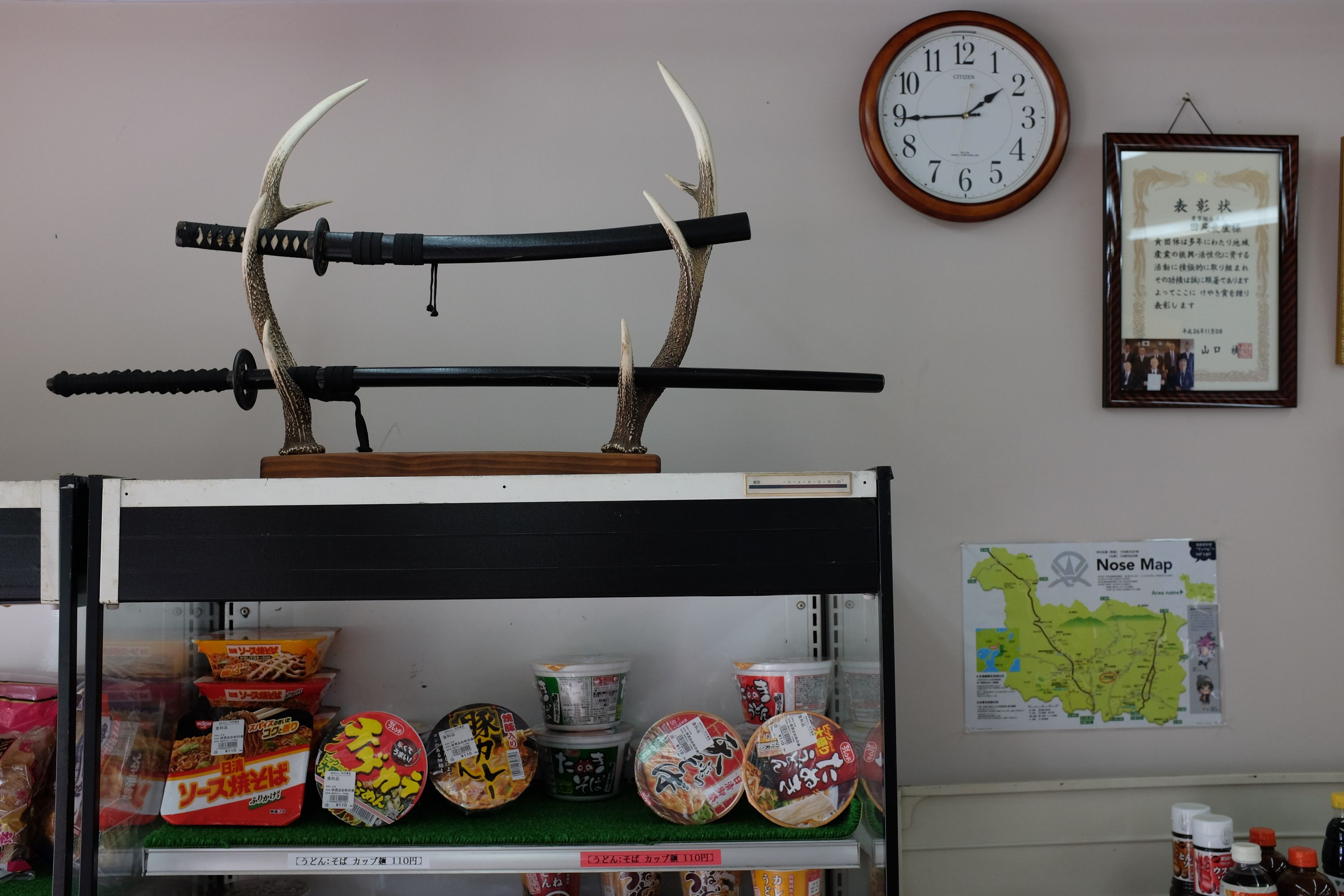 Two Japanese swords displayed on a pair of antlers on the top of a grocery store shelf stacked with cups of instant noodles.