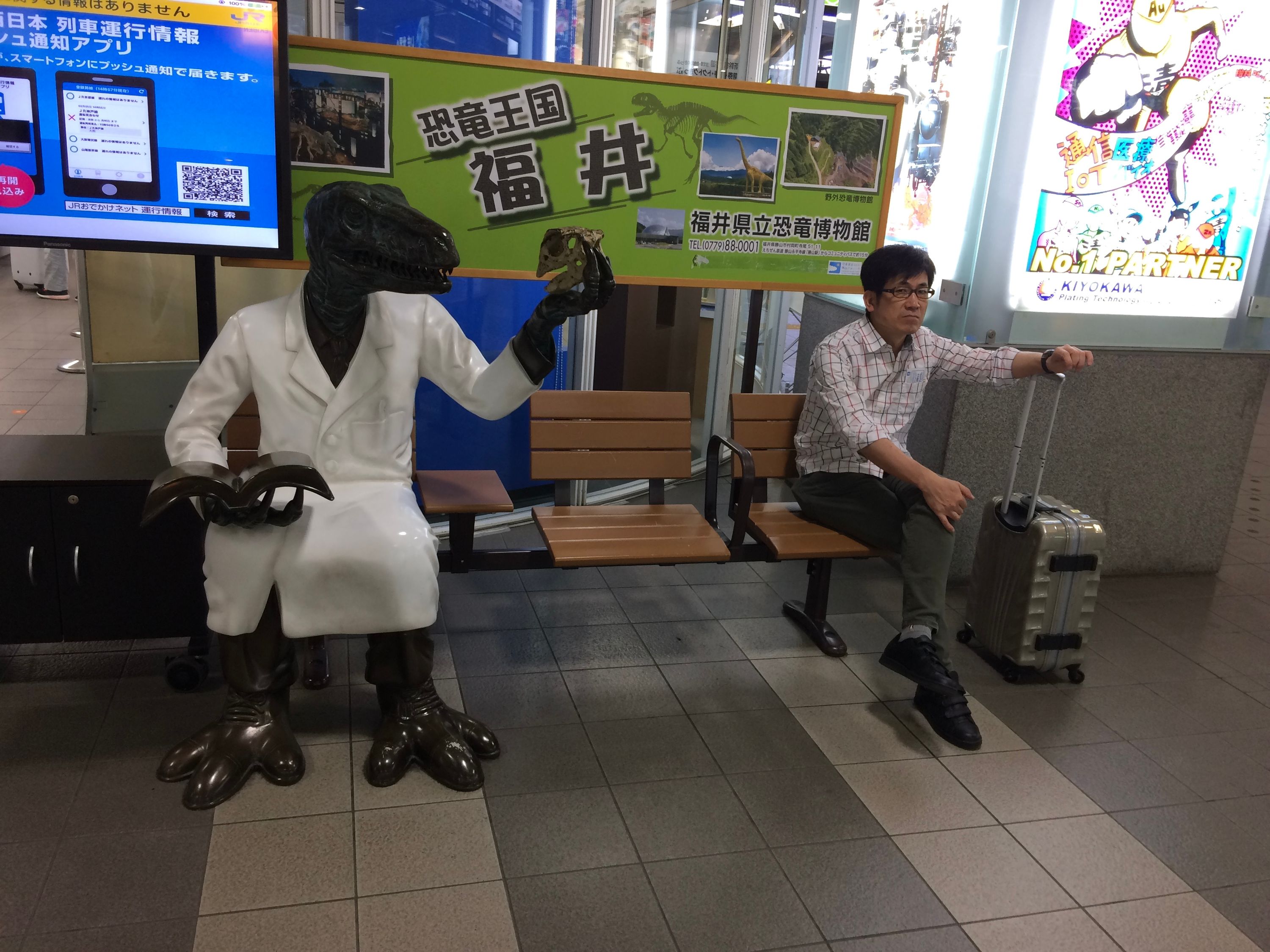 A Deinonychus-like dinosaur, dressed in a lab coat and examining the skull of what appears to be a Fukuisaurus, sits on a bench next to a middle-aged man resting his left hand on a wheeled suitcase.