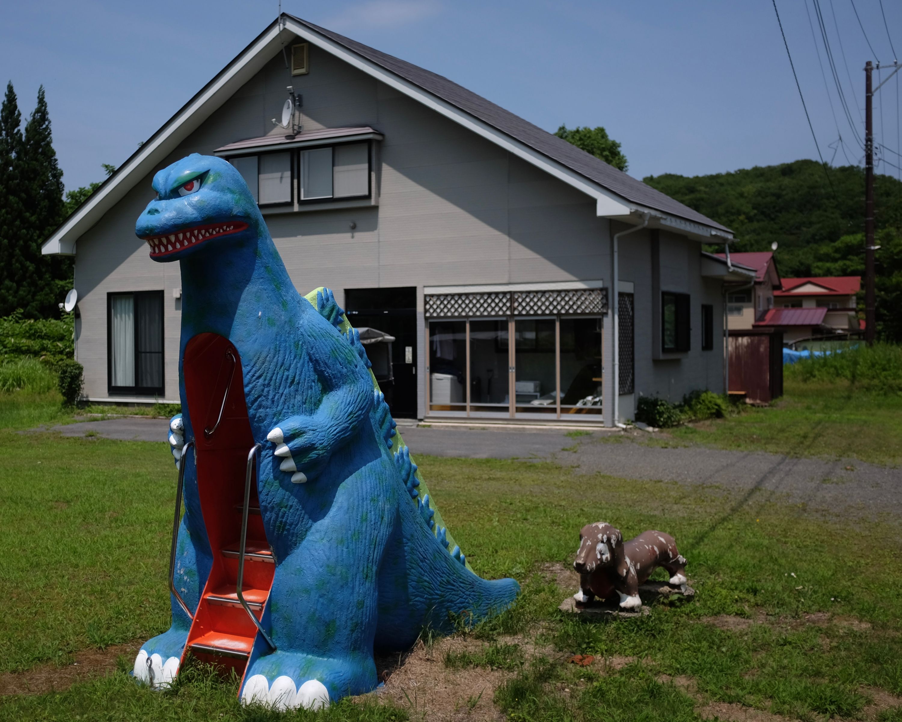 A blue Godzilla-shaped slide and a small brown dachshund stand guard in the front yard of a house.