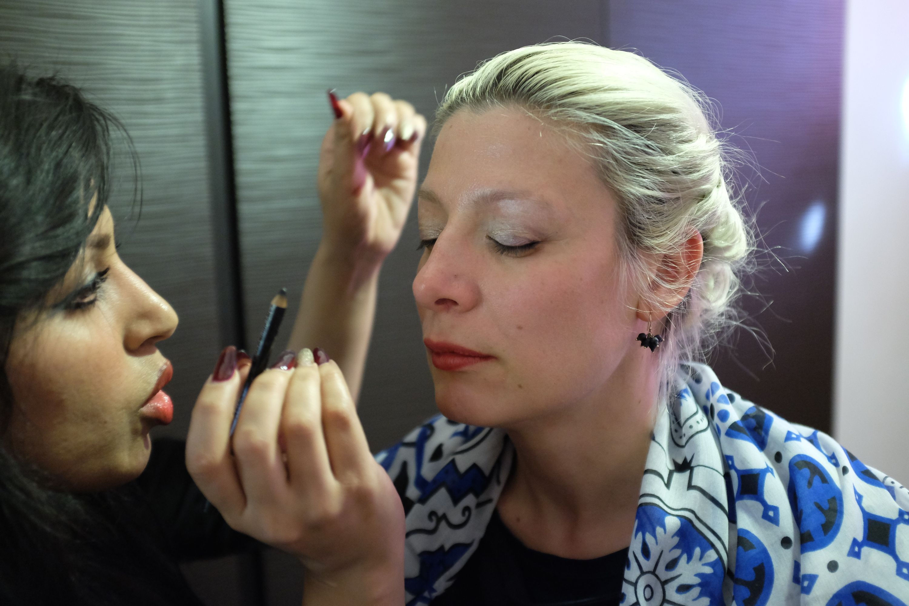 A black-haired woman applies makeup to a blond woman whose eyes are closed.