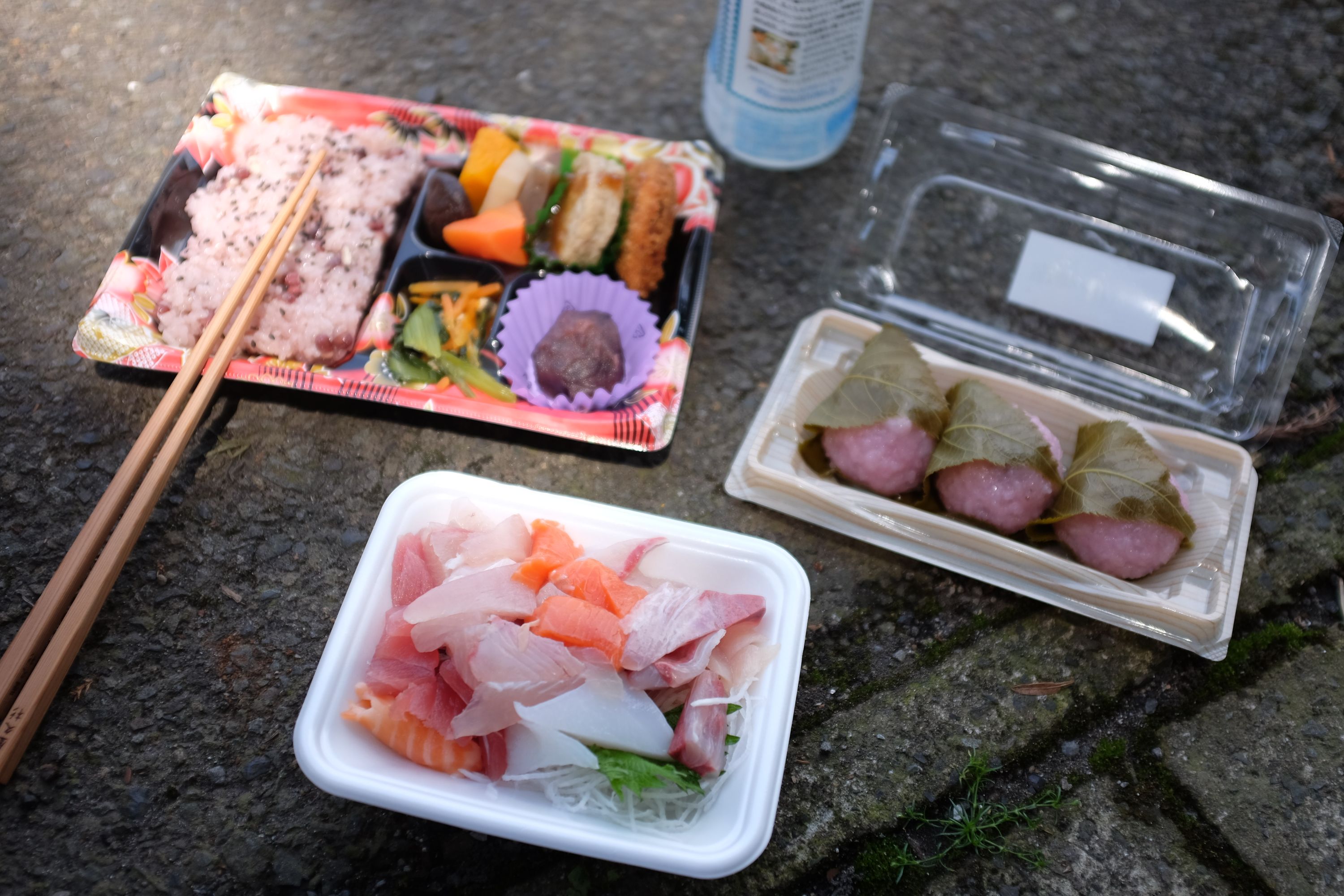 A lunch of sashimi, pink rice balls, and assorted snacks laid on the asphalt in plastic trays.