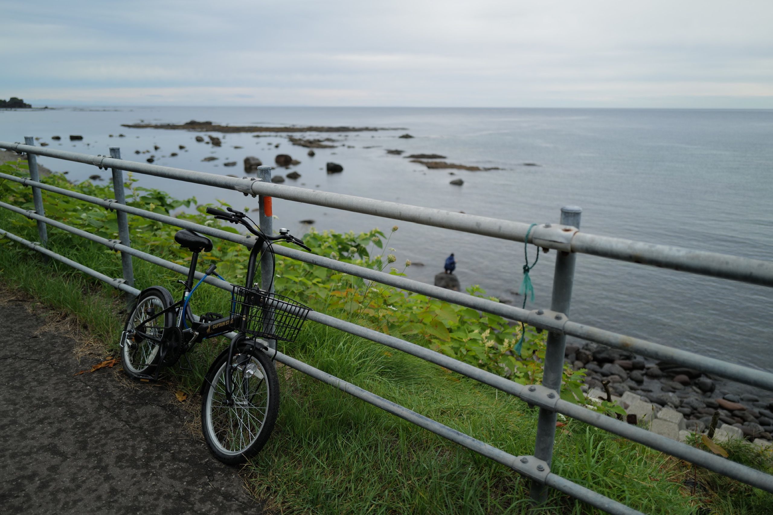 A bicycle leans against a fence by a seashore, with its presumed owner squatting on a rock below