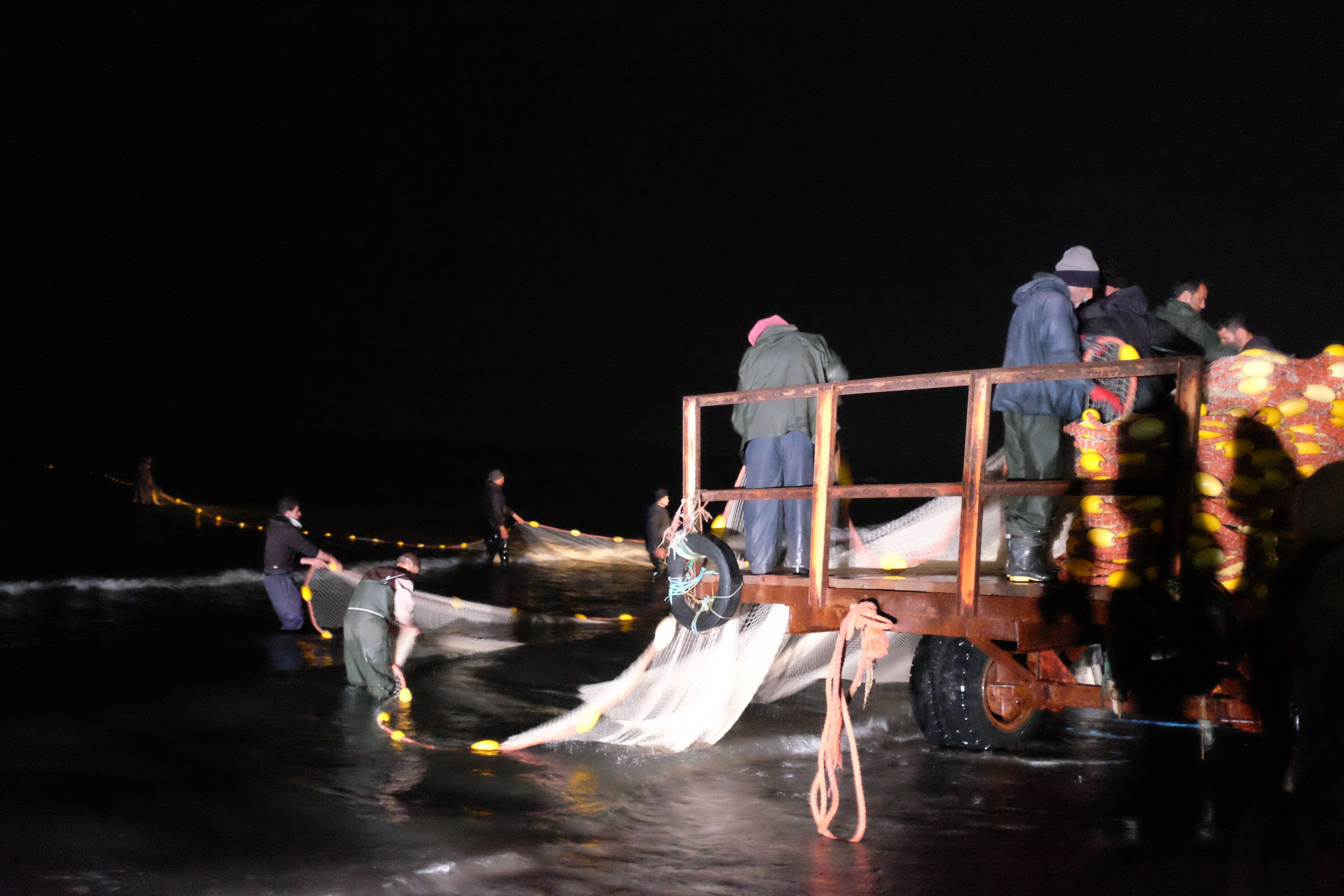 Men drag a fishing net from a sea with a tractor, aided by lights in the complete darkness.