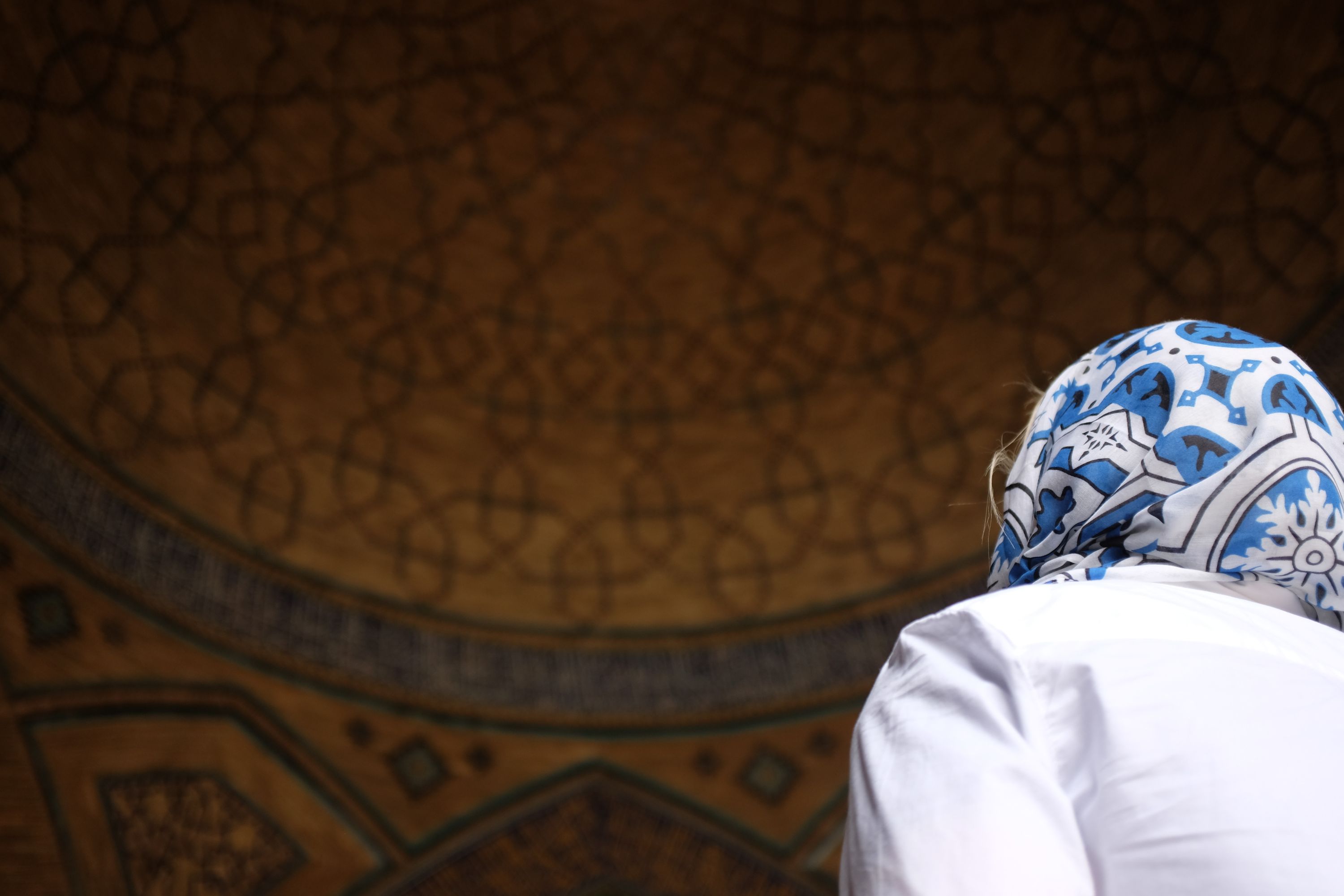 A woman in a blue headscarf and a white shirt looks up at the intricately patterned ceiling of a mosque.