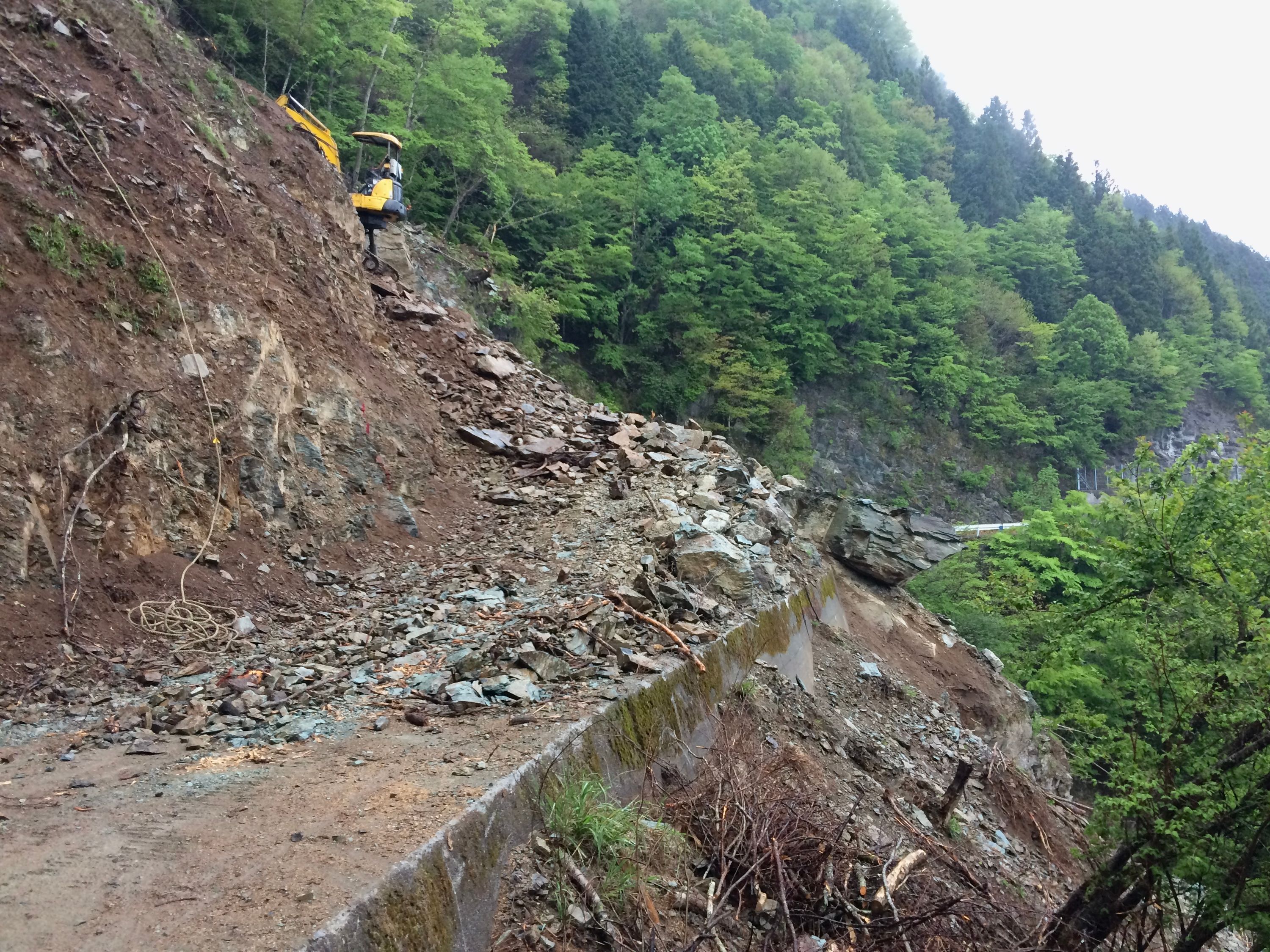 A mountain road completely covered by rocks from a landslide, making it impassable to vehicular traffic. A very small yellow excavator is parked on the rocks.