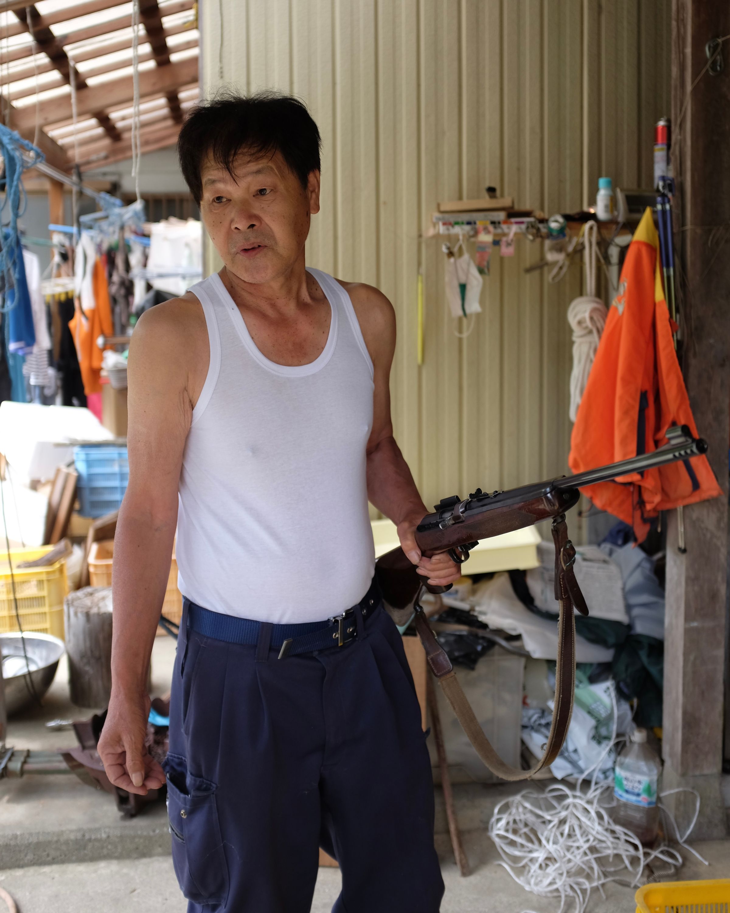 The same Japanese man, dressed in a sleeveless t-shirt, holds a rifle in his left hand while looking off-camera.