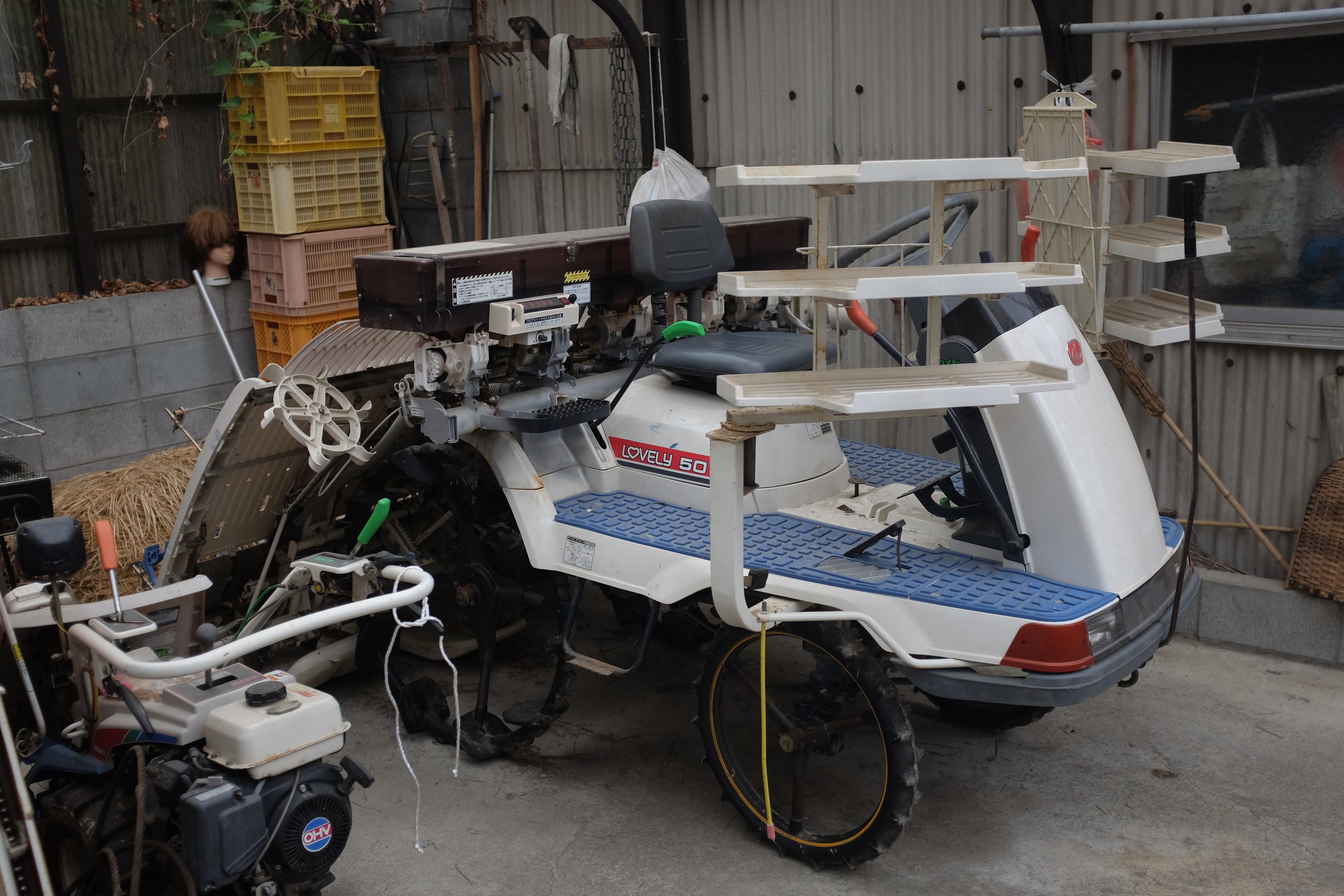 A small rice planting tractor parked in a garage with a mannequin’s head on a shelf behind it.