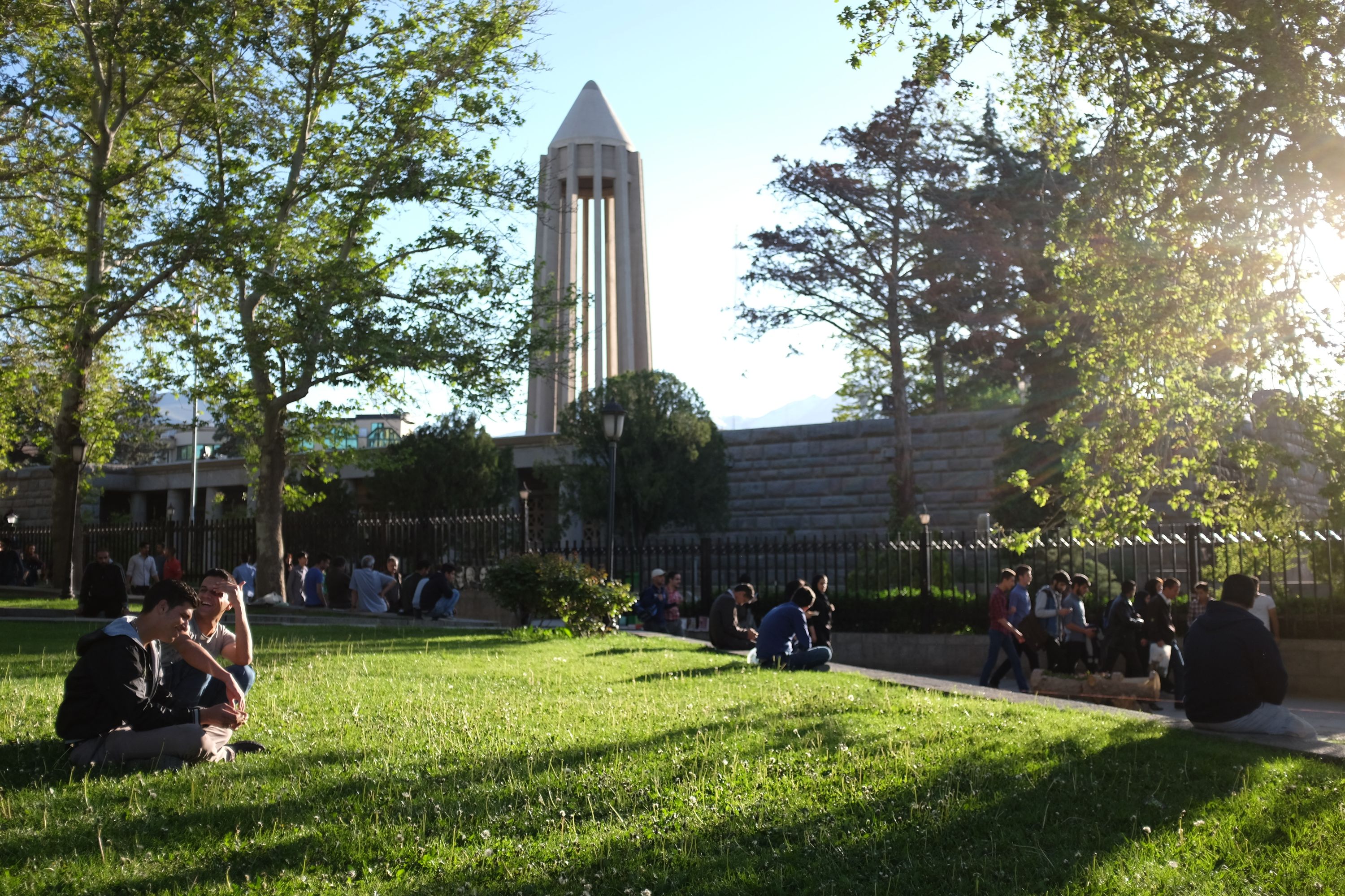 People sit in a park in the evening light in front of a tall building which looks like a concrete rocket.
