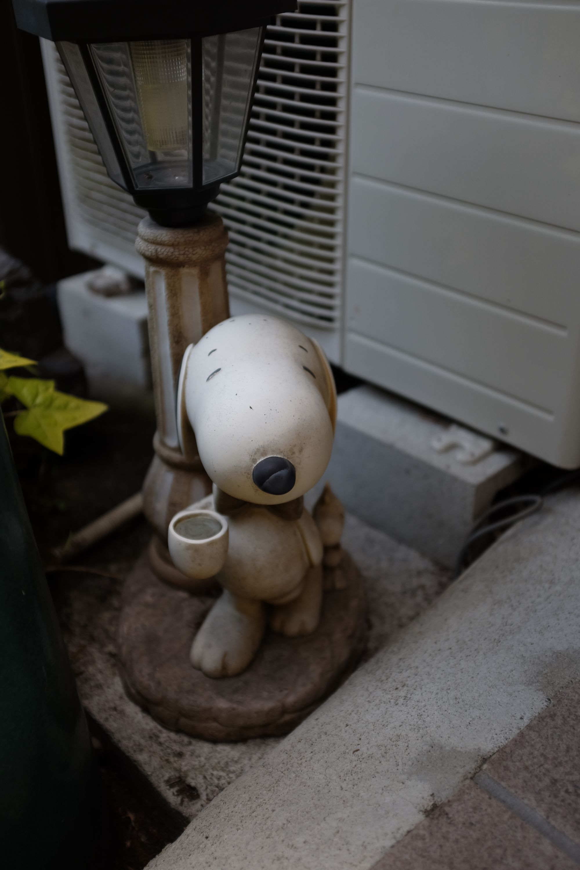 A Snoopy figure holds a coffee cup by a lantern in front of a house.