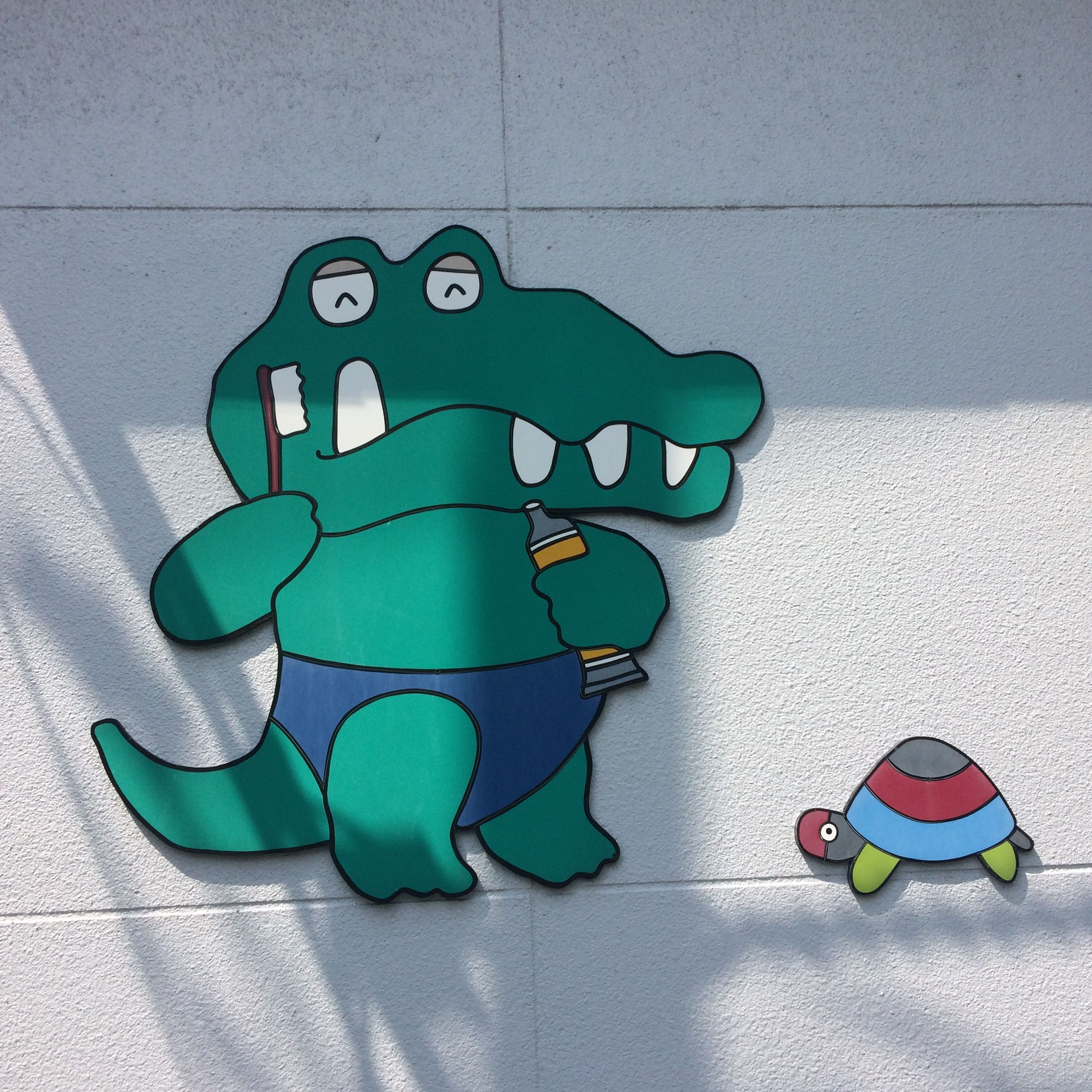 On a wall, a cartoon alligator brushes its teeth while a small tortoise looks on.