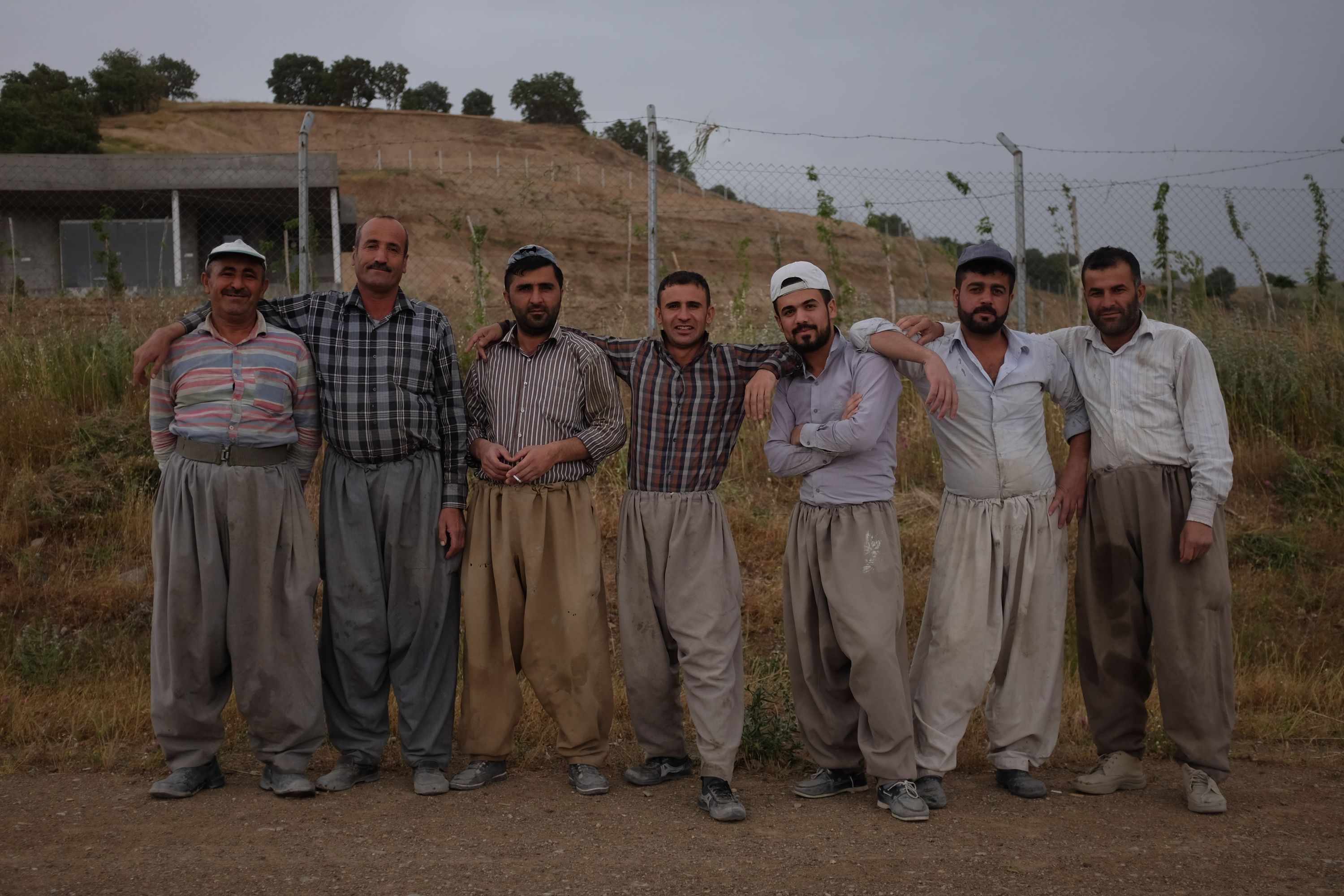 Seven men, all dressed in wide-legged Kurdish trousers, stand in a line by a fence.