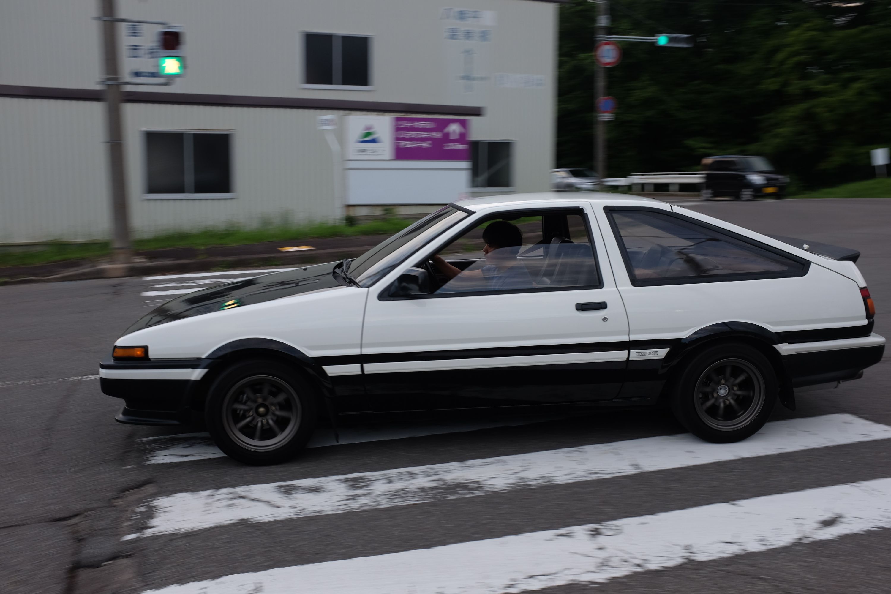 A Hachi-Roku, a black and white Toyota AE86 sports car, takes a turn across a pedestrian crossing.