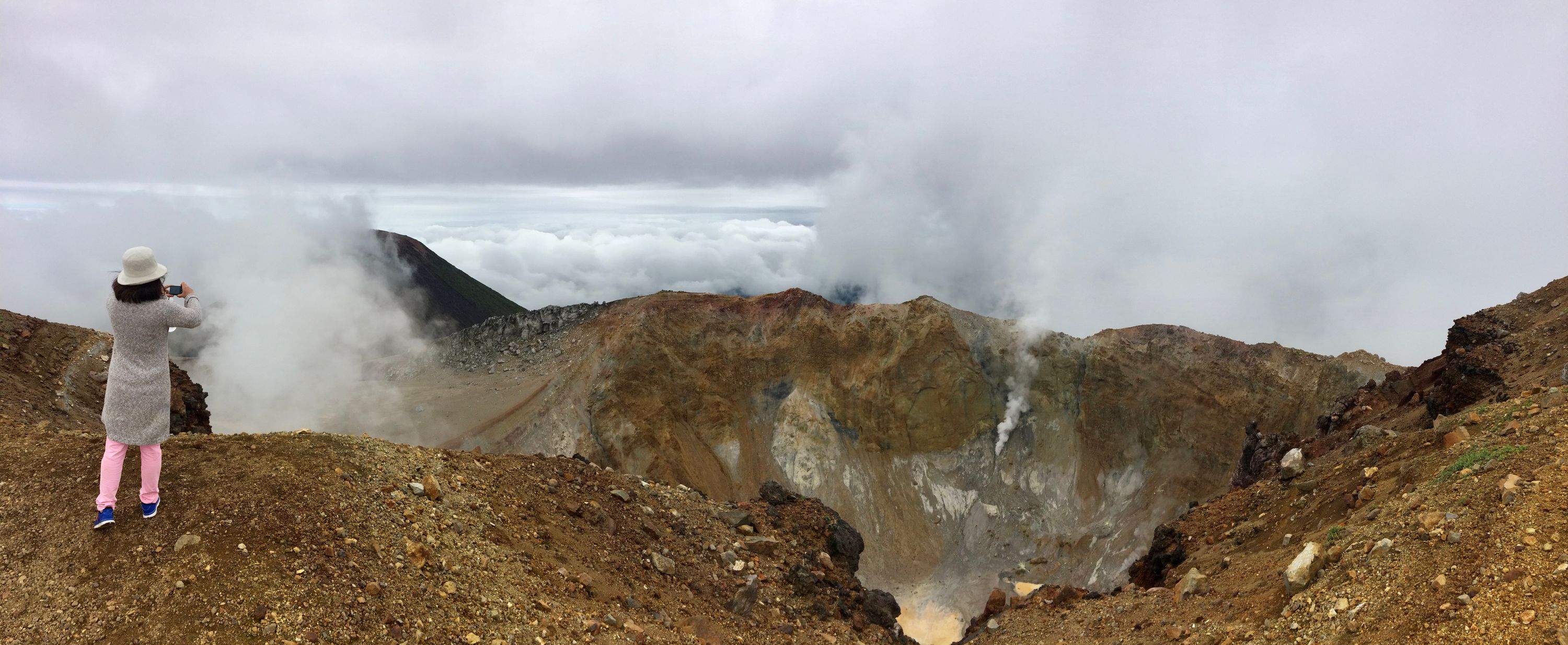 Panorama of a woman in pink trousers and a grey cardigan photographing the volcanic crater.