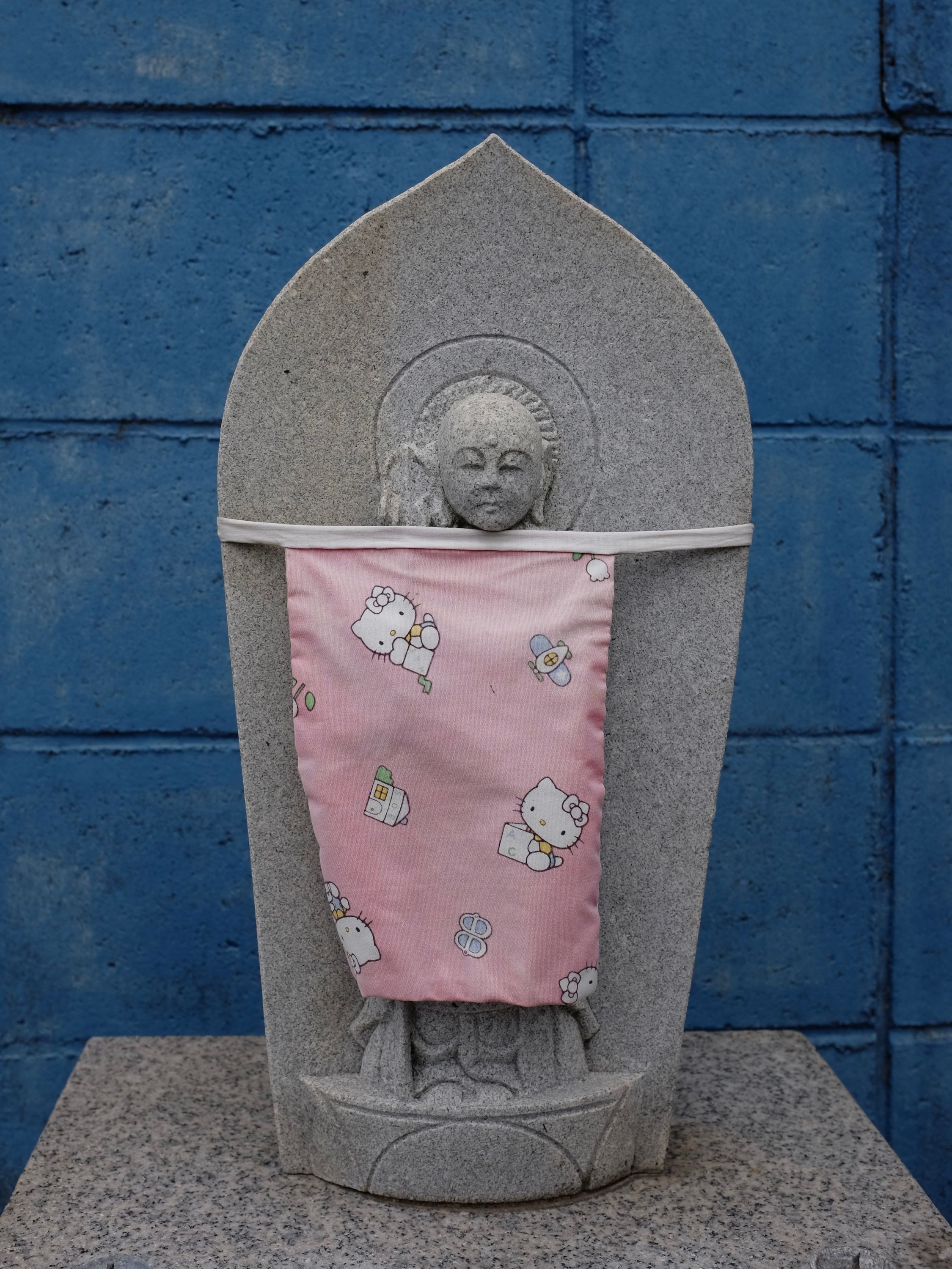 A Buddhist statue wearing a pink apron with a Hello Kitty pattern stands against a blue wall.
