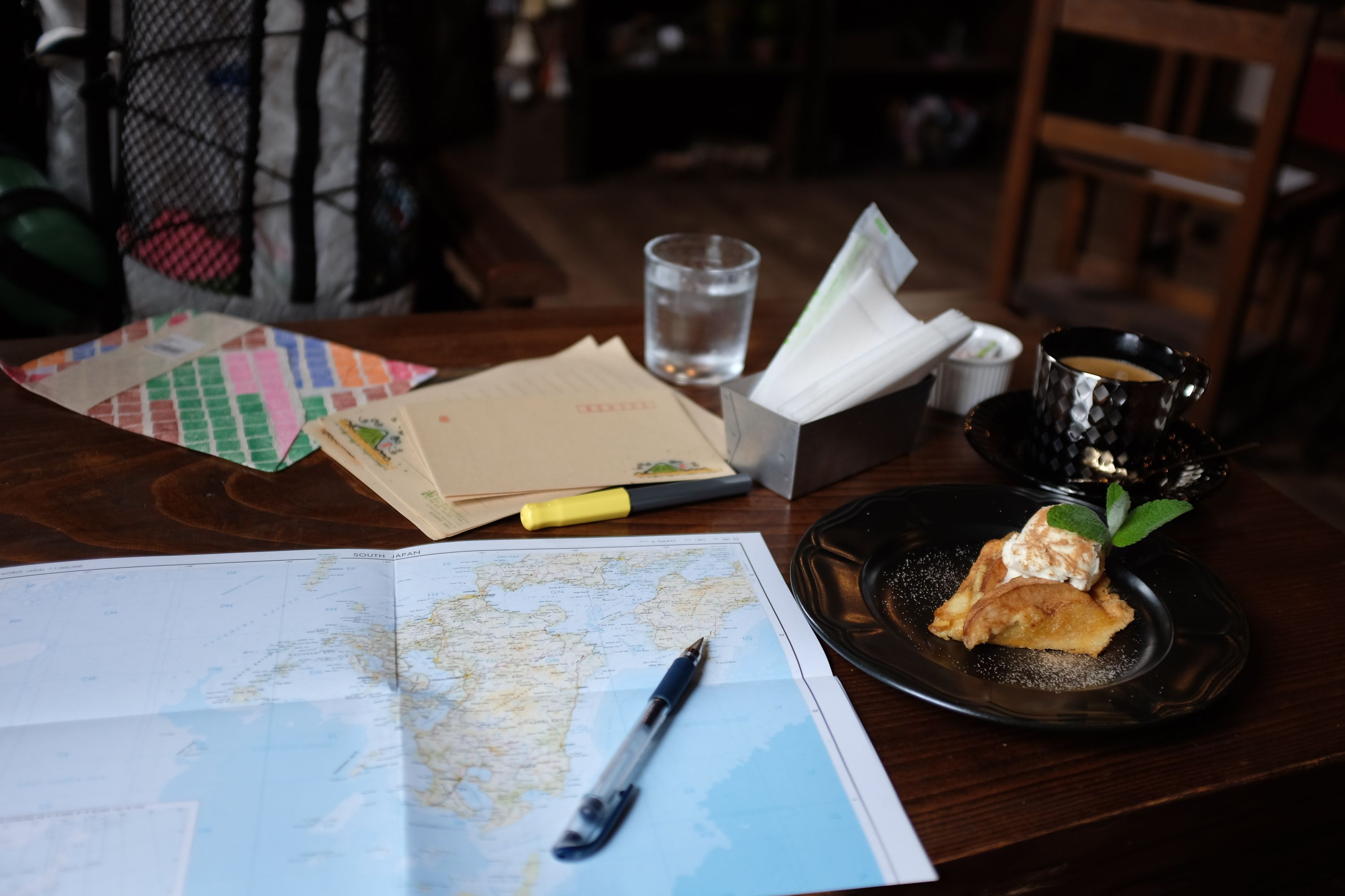 A map of Kyushu laid out on a coffee table, with some pens, envelopes, a cake, and a cup of coffee.
