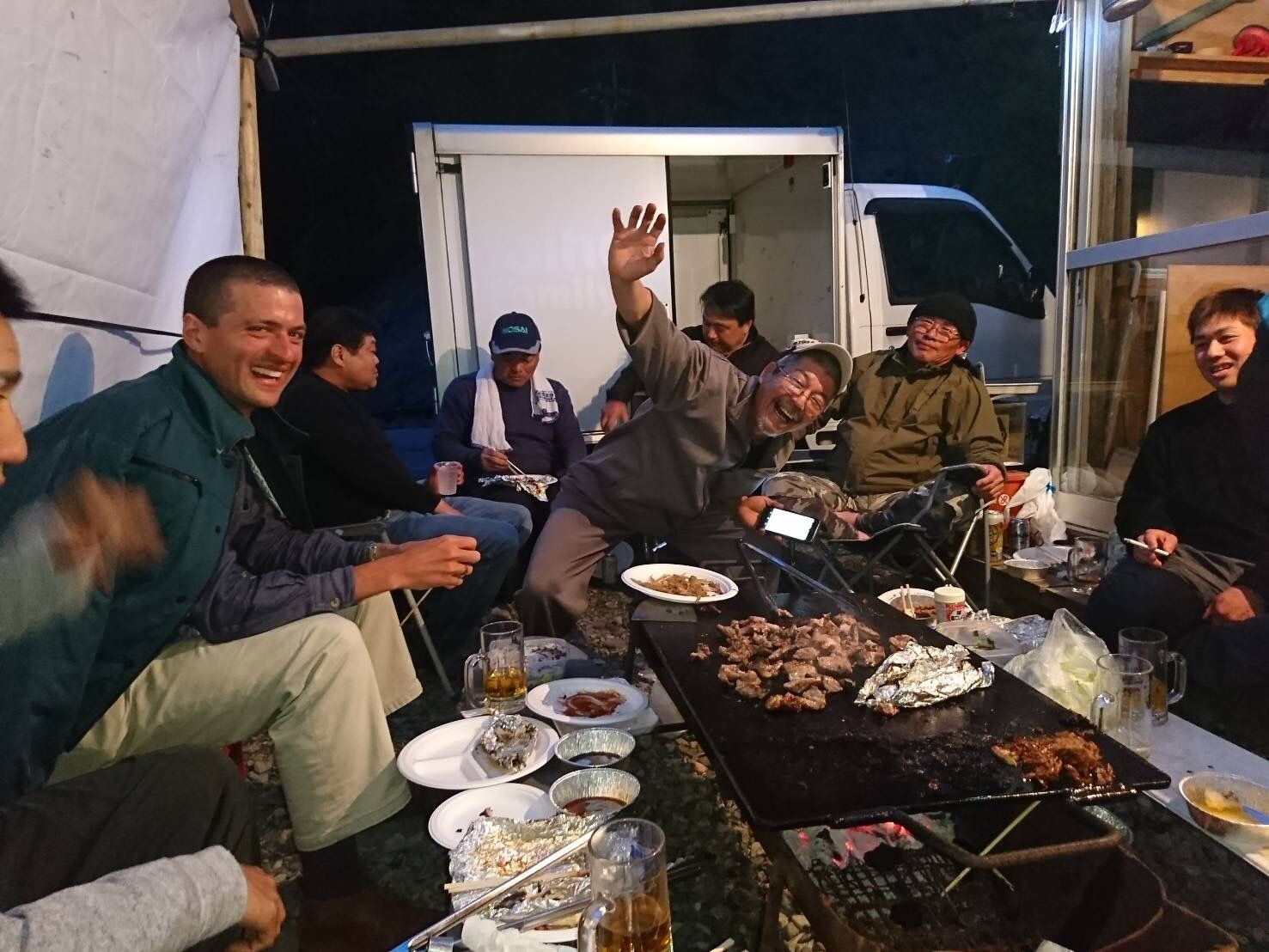 The author with a large group of men sitting around a huge grill with a lot of food, all of them clearly enjoying themselves.