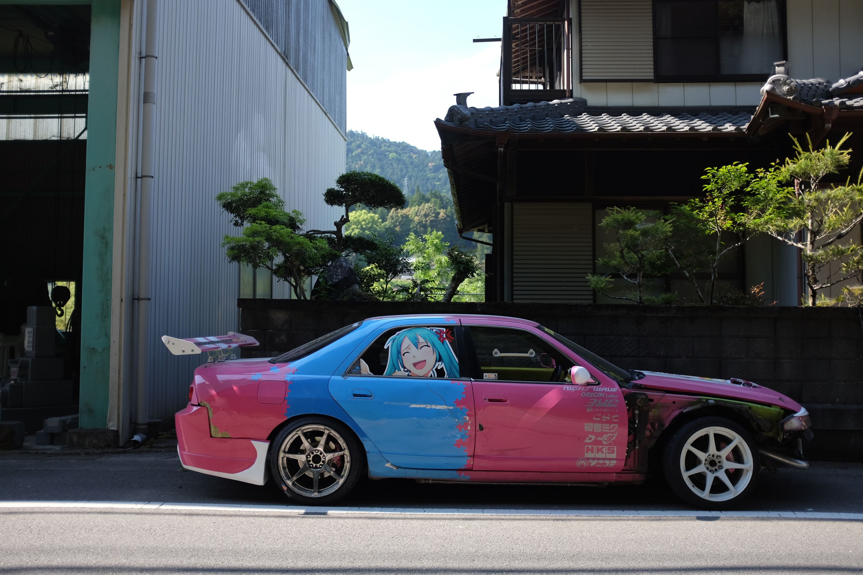 A pink and sky blue Nissan Skyline R32 parked in front of a Japanese house.
