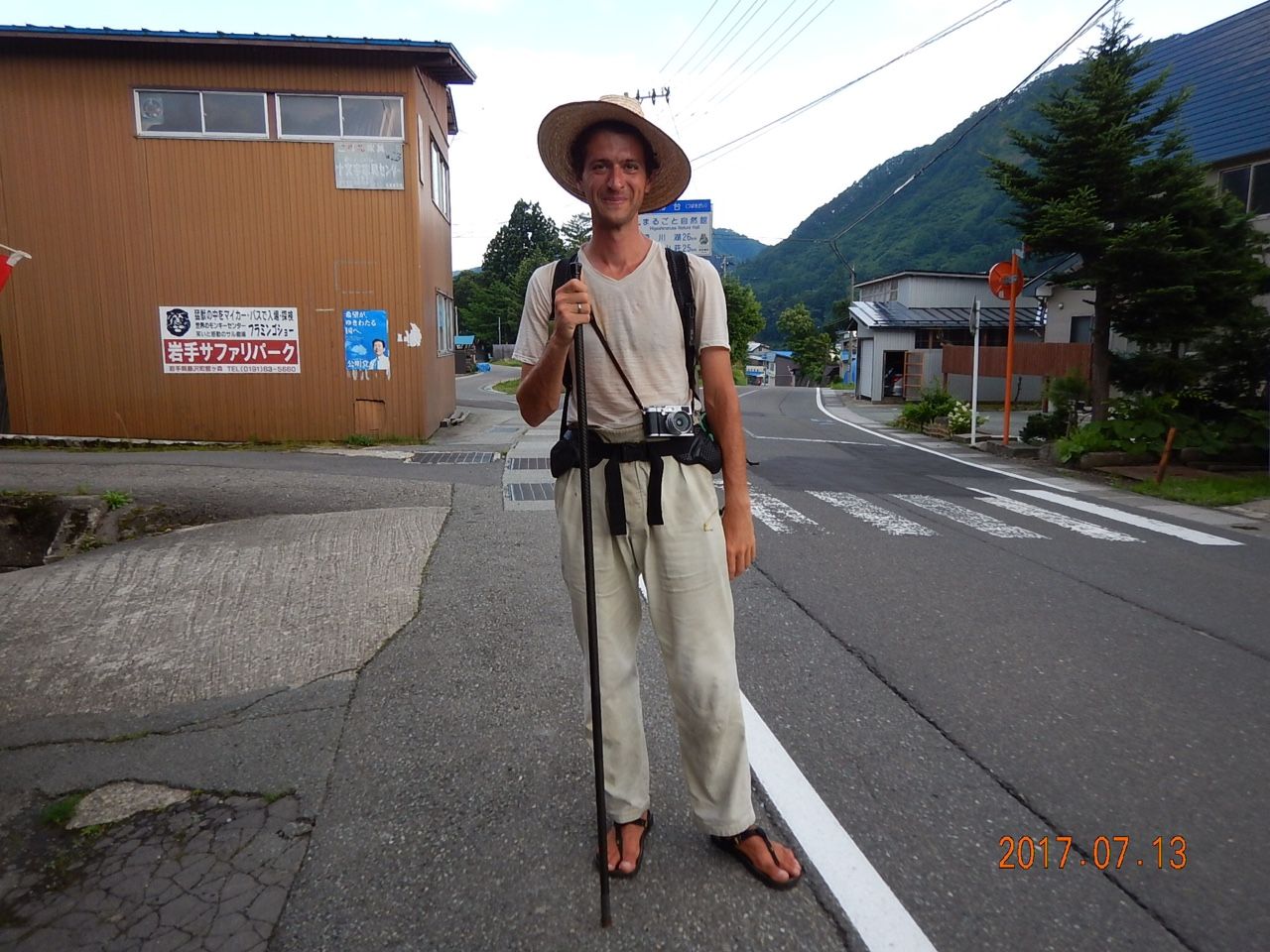 Peter Orosz, in a straw hat and holding his walking stick, stands by the side of the road.