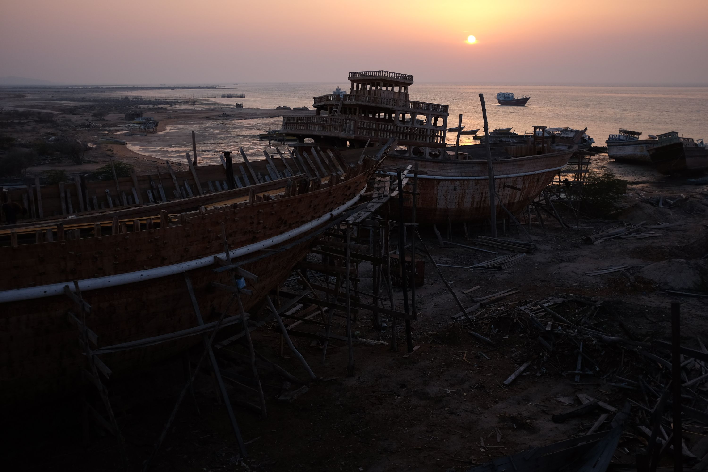 Two Arabian-looking boats under construction on the shore, with a completed one floating by in the sunset.