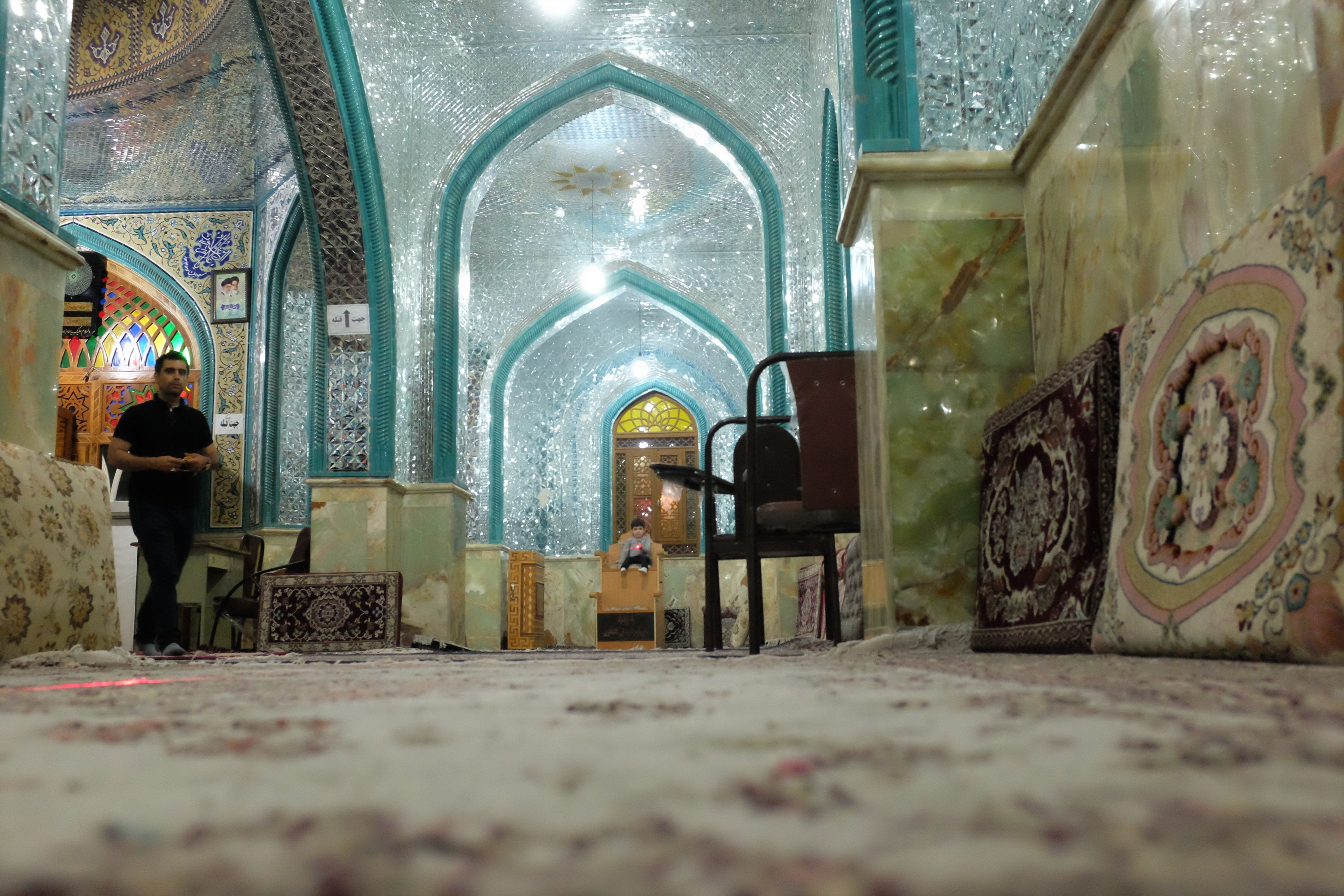 Seen from ground level, a small boy sits on a tall chair and plays with a laser pointer in a shrine whose ceilings are decorated entirely with mirrored tiles.