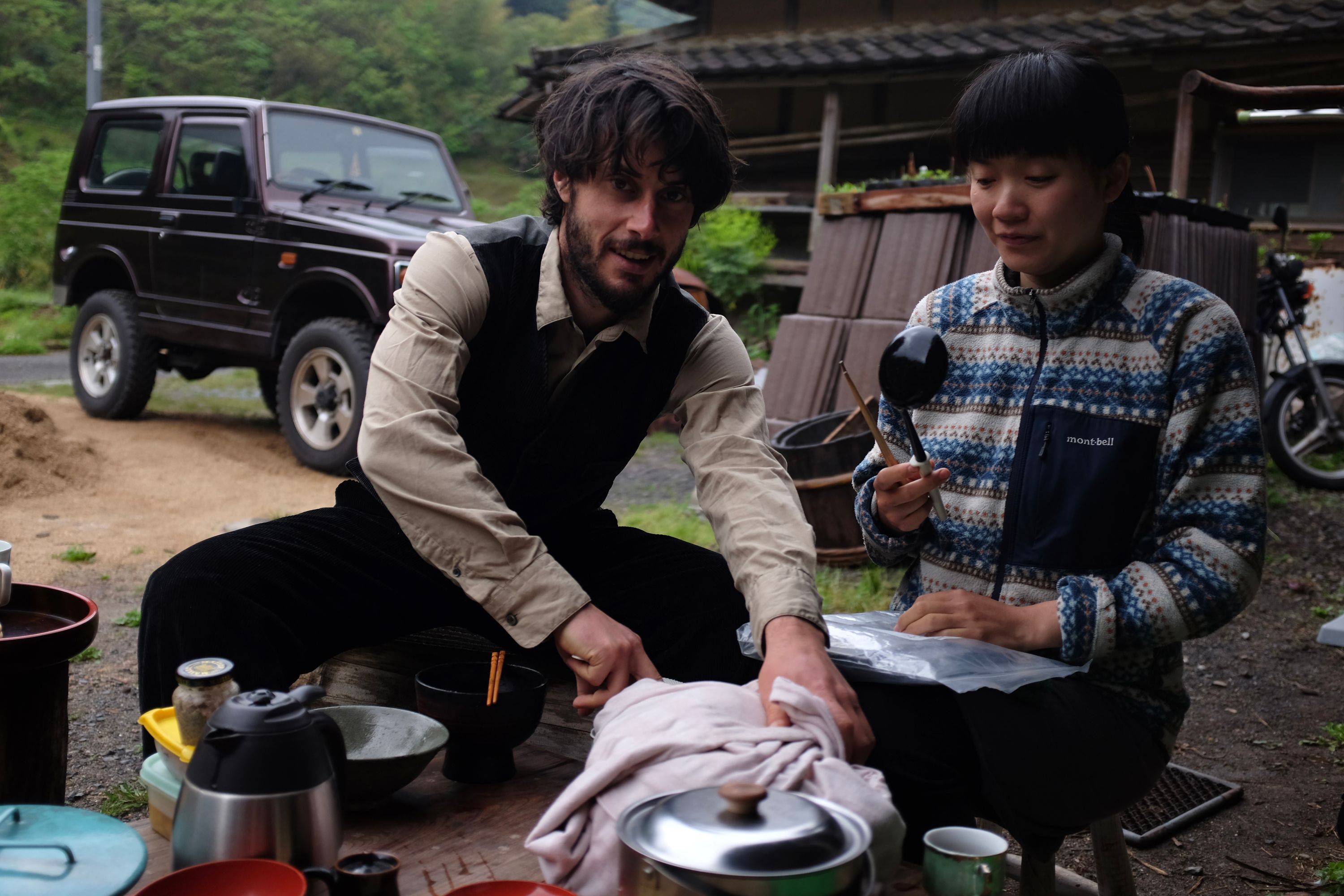 A French man in a shirt and a waistcoat and a Japanese woman in a cardigan sit outside their house and prepare to eat breakfast.