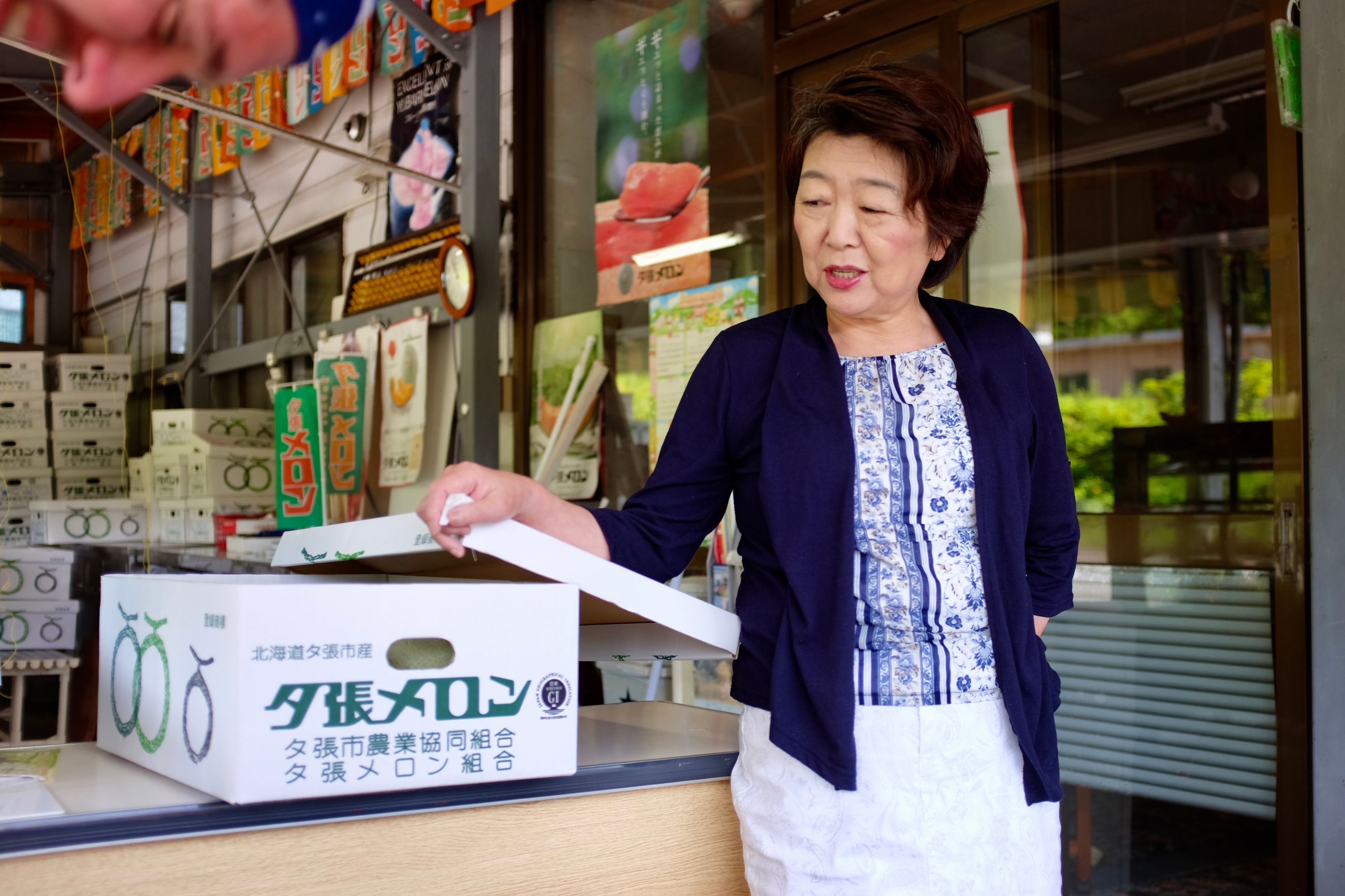 A woman selling melons looks at a crate labelled “Yūbari Melon”.