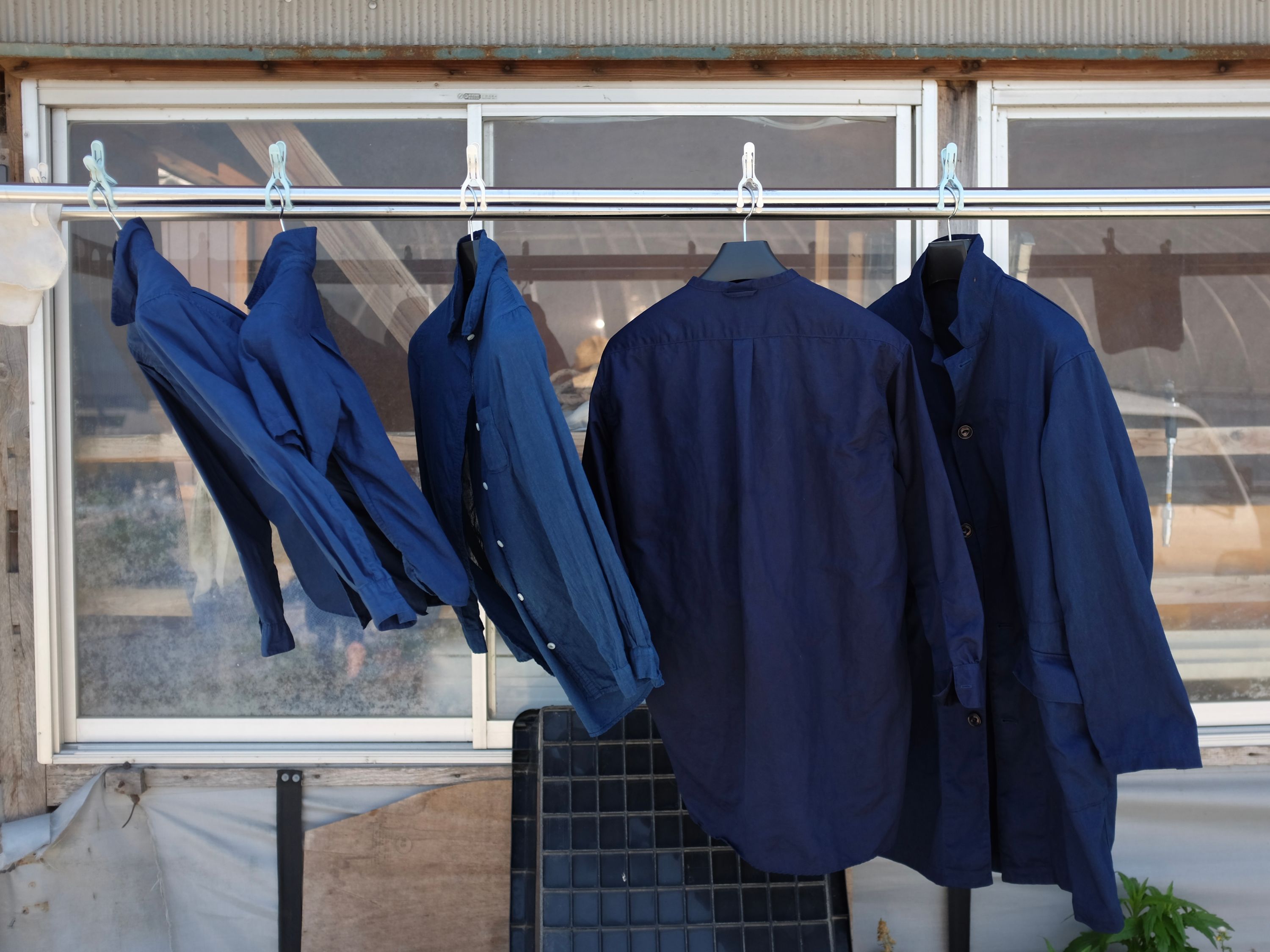 Three shirts and two jackets, all of them dyed indigo, drying in the breeze on a rail.