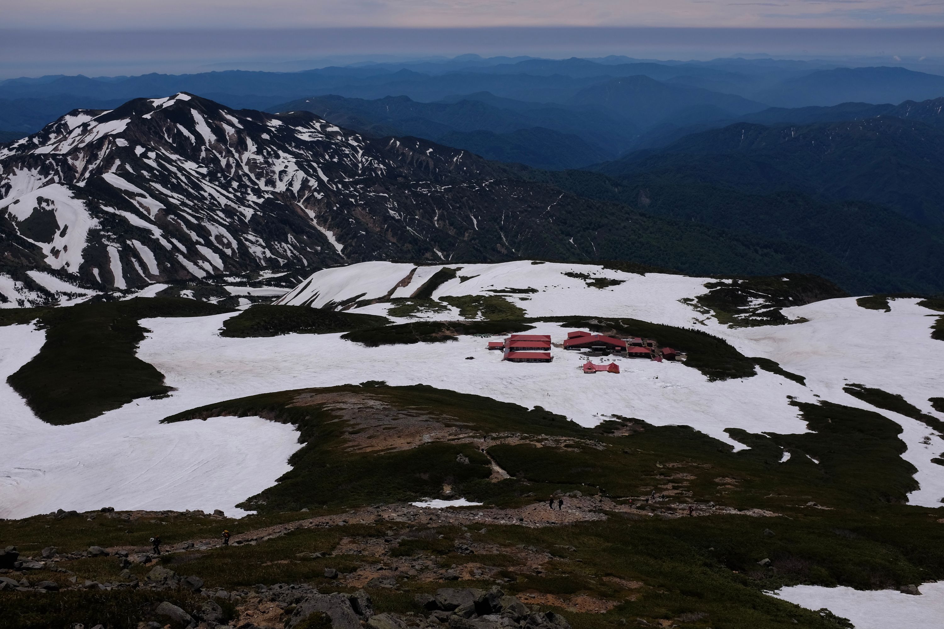 Looking down on a cluster of red-roofed mountain huts in a snowfield, behind them is a range of lower mountains.
