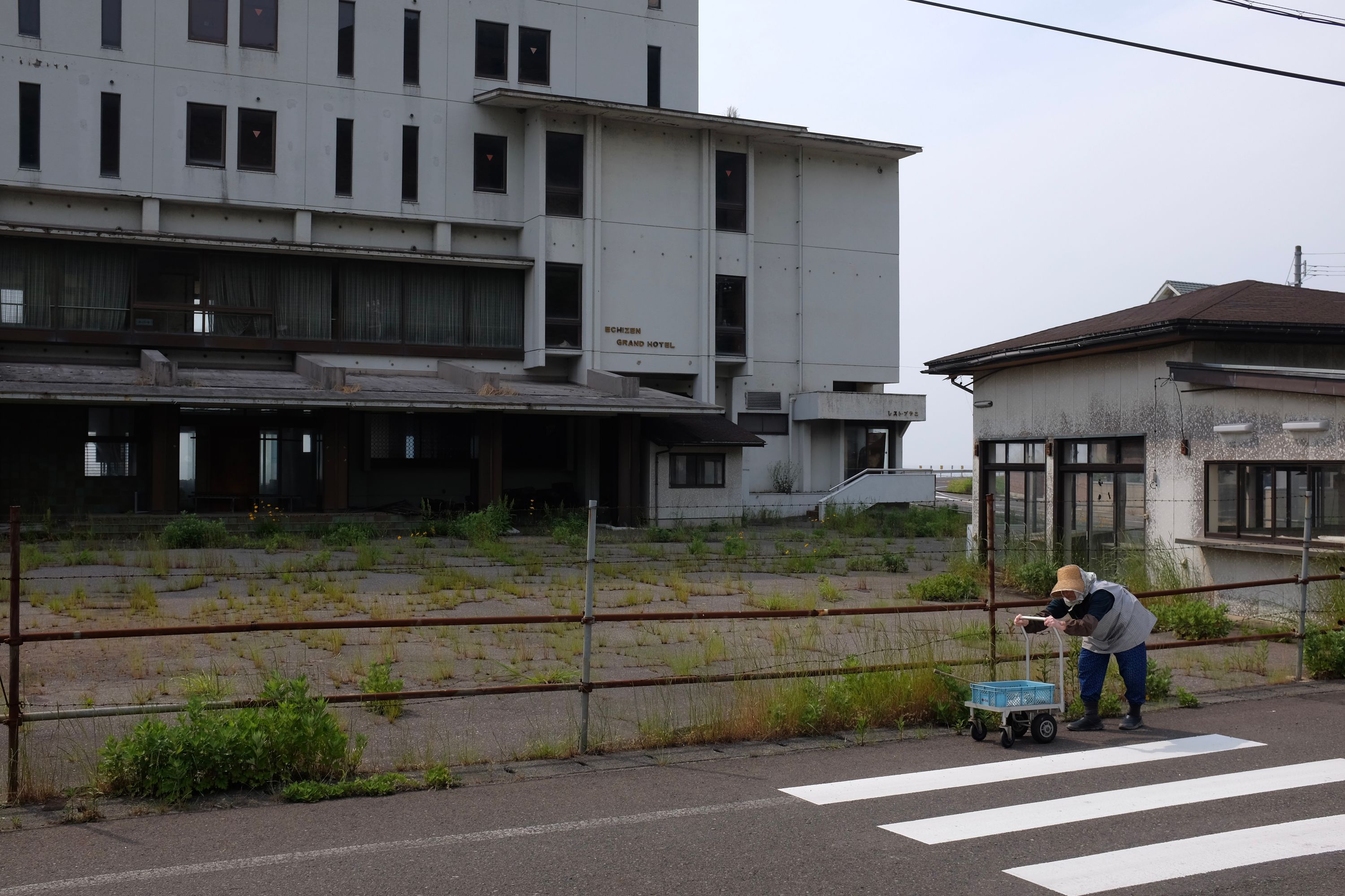 An old woman pushes a wheeled walker in front of an abandoned building marked “Echizen Grand Hotel”.