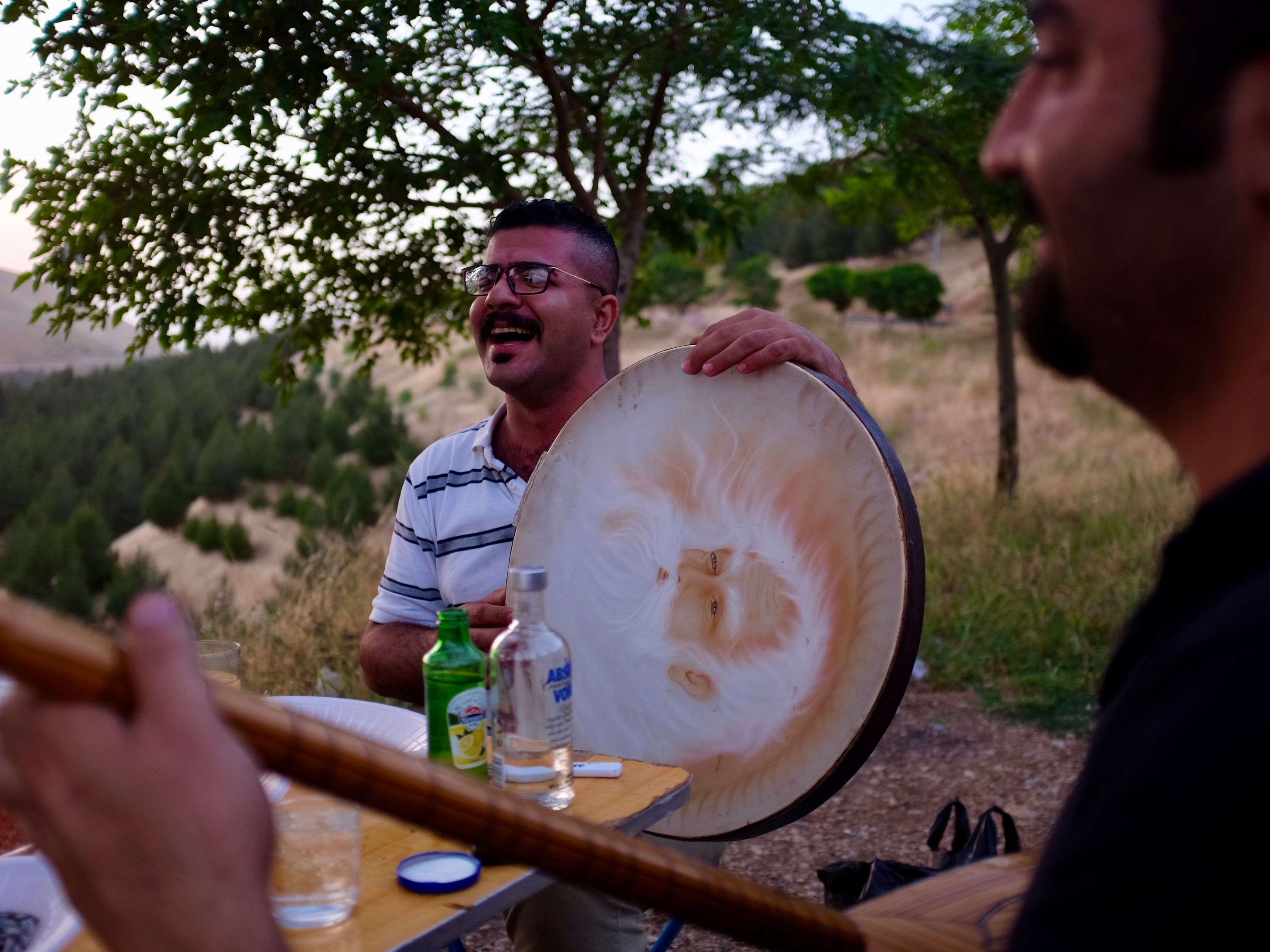 A man plays a large, handheld drum, and another a guitar-like instrument at a table on a hillside in the evening light.