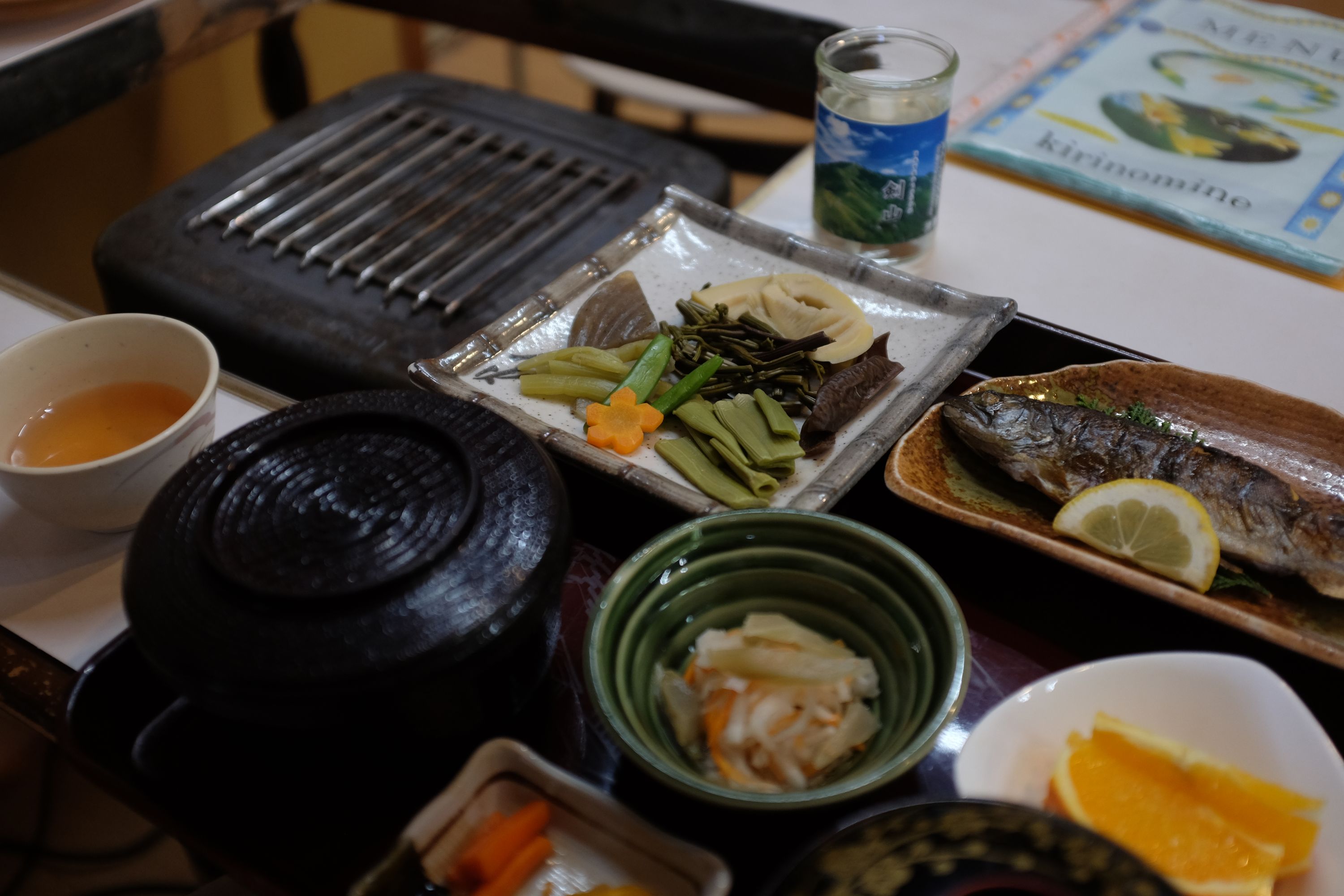 A lunch menu of vegetables, grilled fish, fruit, tea, and sake arranged on a table.