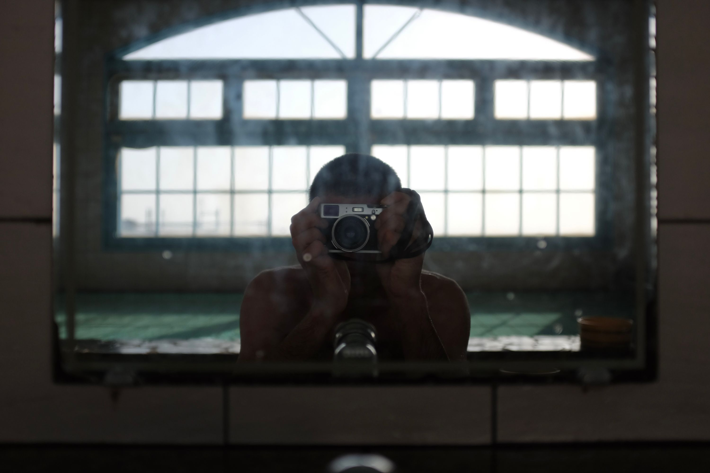 The author takes a self-portrait in the mirror of a public bath, the mirror obscuring his face, a large window visible behind him.