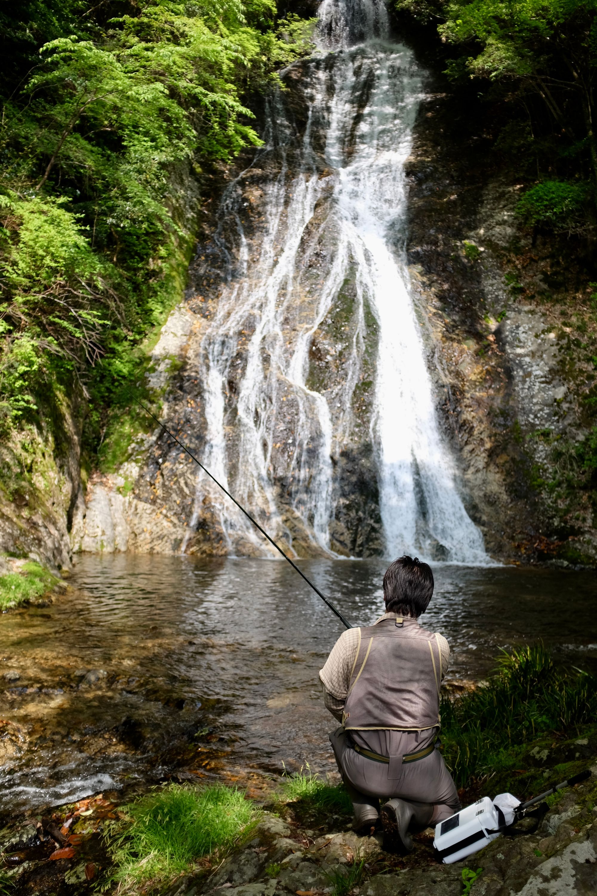 A man fly-fishes in the pool of a high waterfall directly across from him.