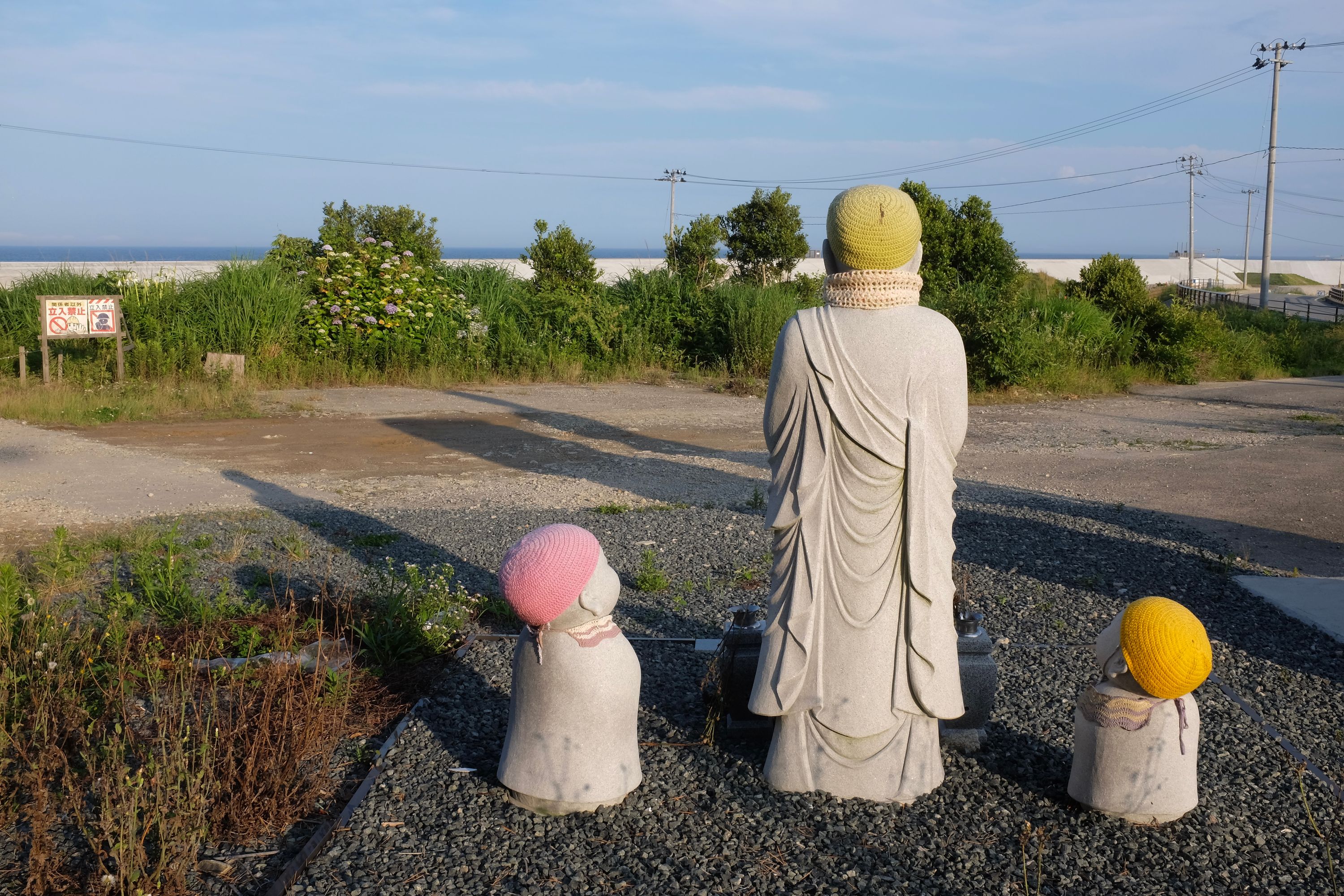 Three Buddhist statues in knit caps facing towards the ocean.