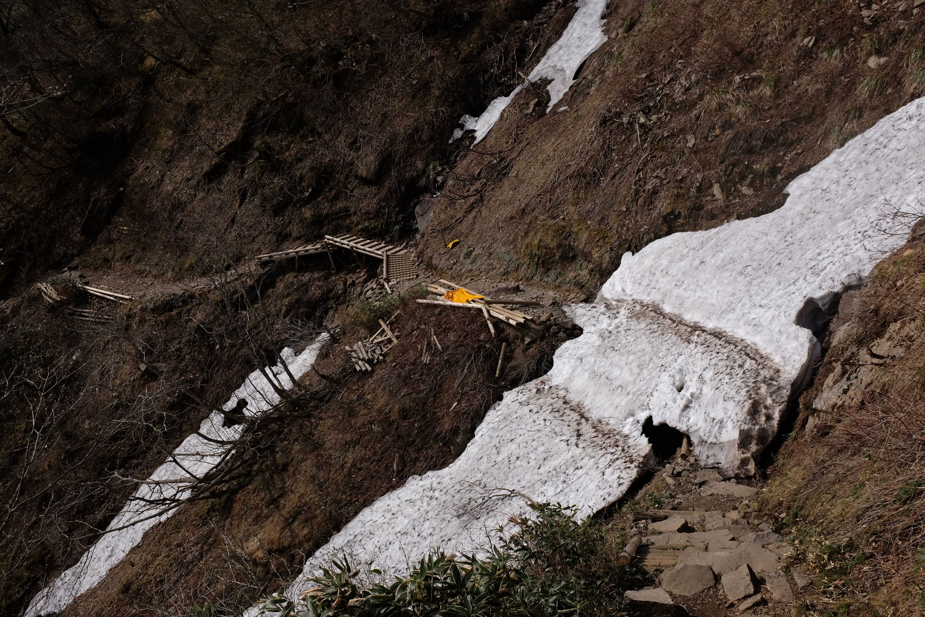 Two lingering patches of snow cut across a trail traversing a steep hillside, a broken wood bridge between them.