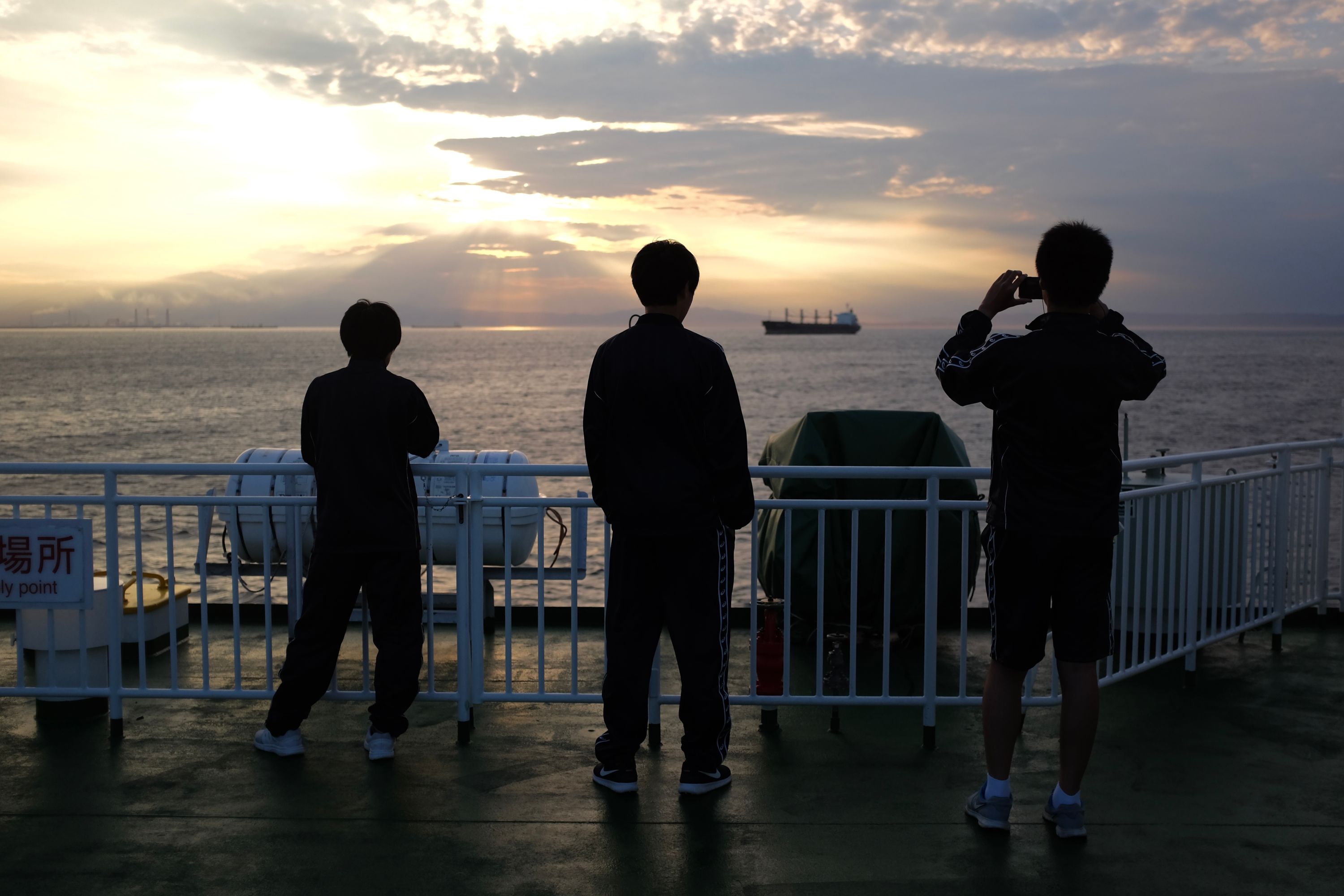 Three boys on the deck of the ferry look at the setting sun, one of the taking a picture with his phone.