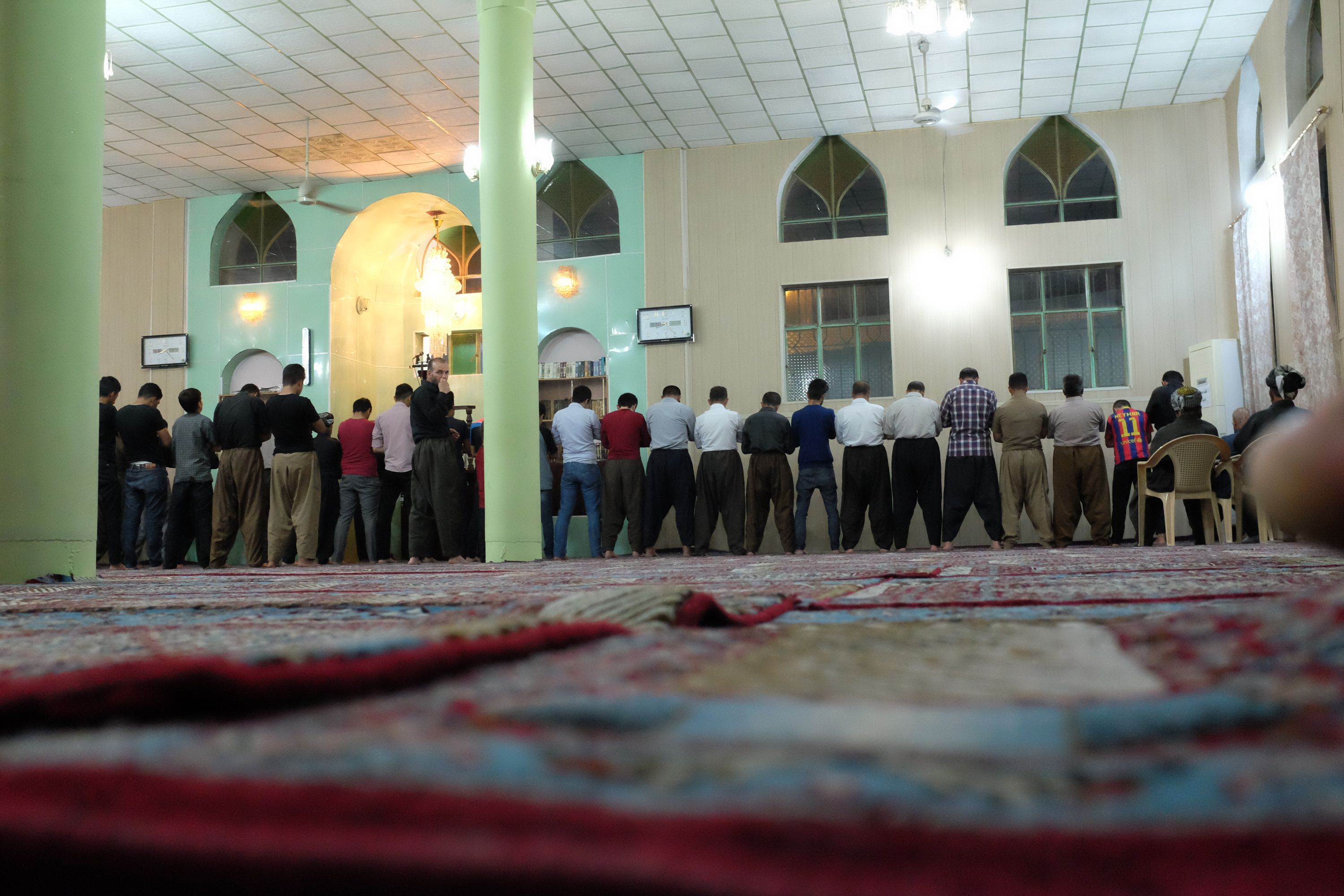 A line of men stand in a mosque during prayer, all but one facing away from the camera.
