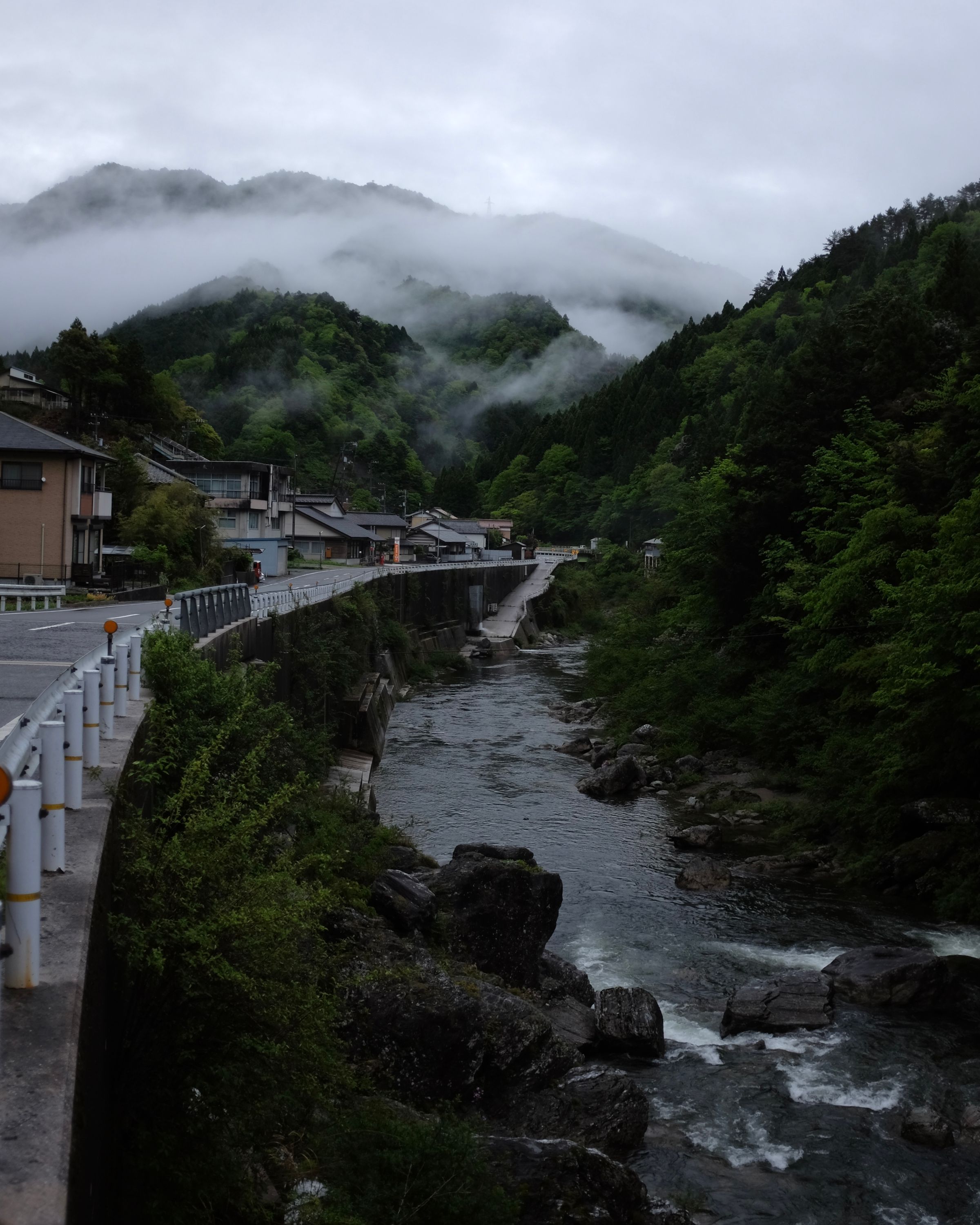 A mountain river runs by a village road, with misty, forested mountains in the background.