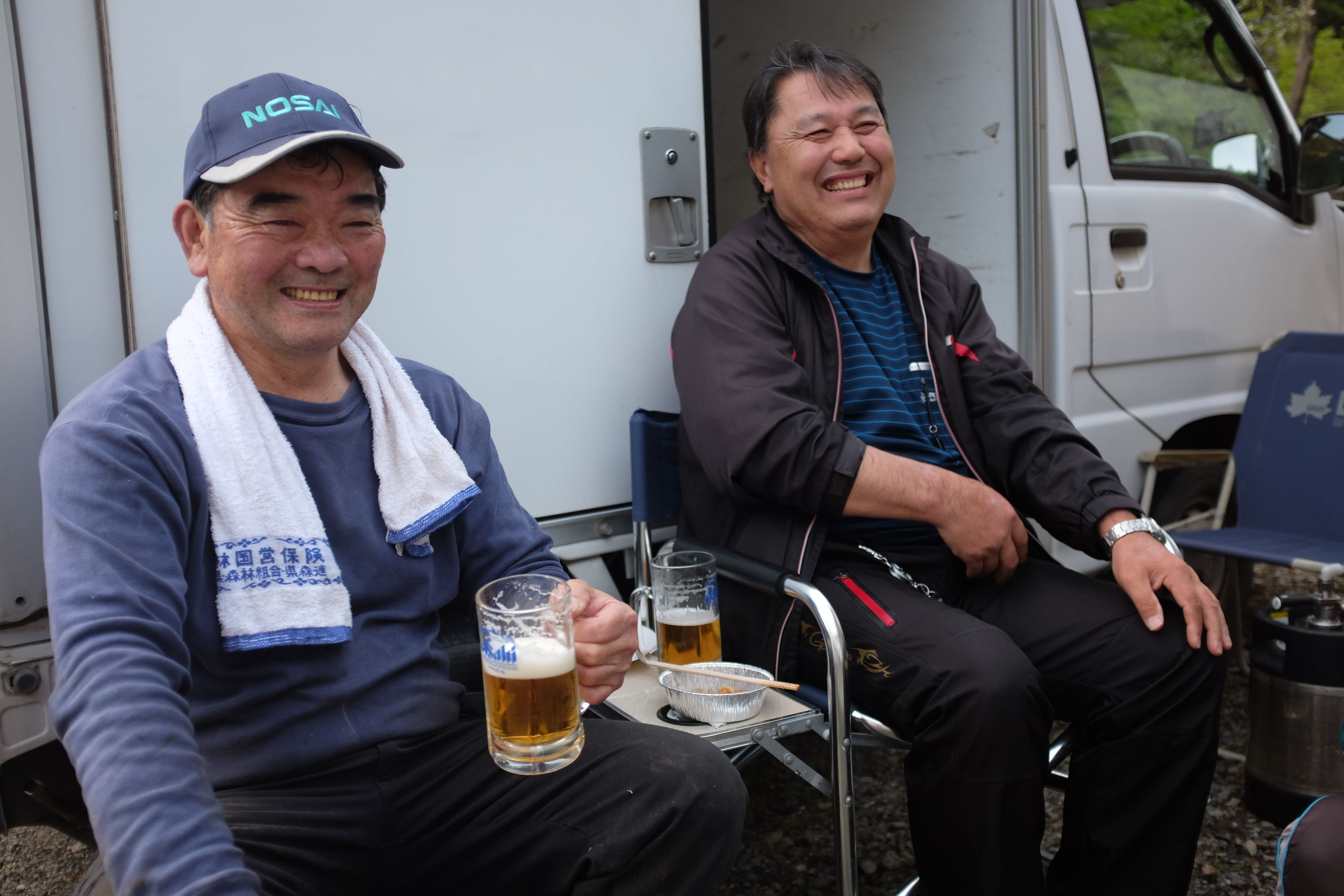 Two Japanese men sit in camping chairs by a truck and enjoy large glasses of beer.
