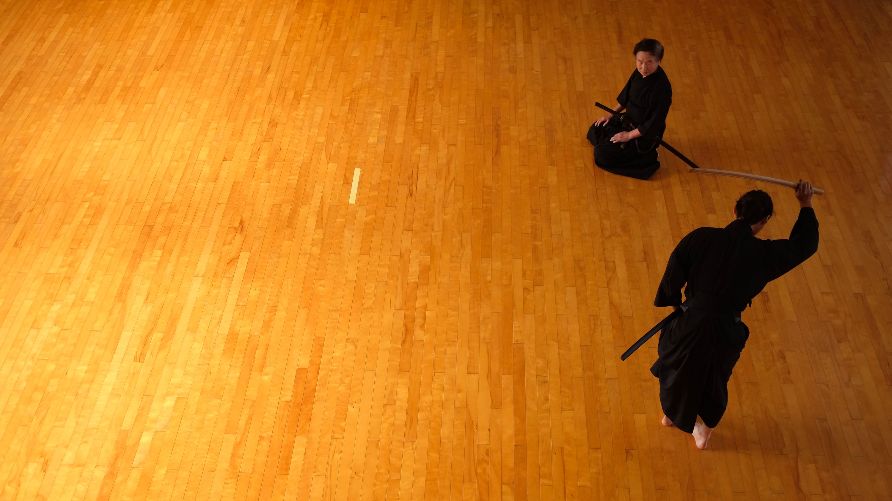 Two Japanese men dressed in hakama, Japanese martial arts clothes, train with Japanese swords on a parquet floor.