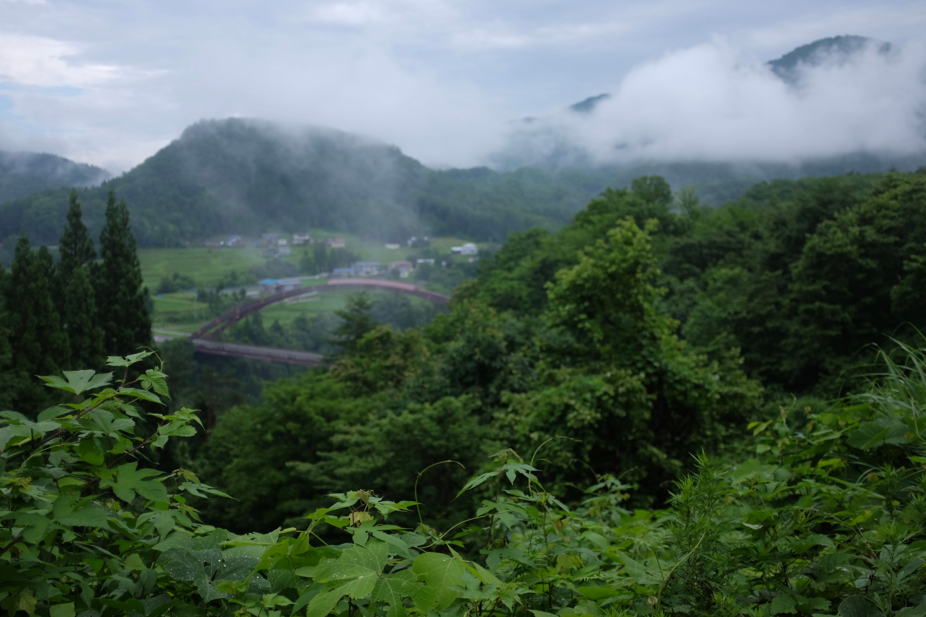 Panorama of a hilly, very green, cloudy landscape, with a bridge and a village in the medium distance.