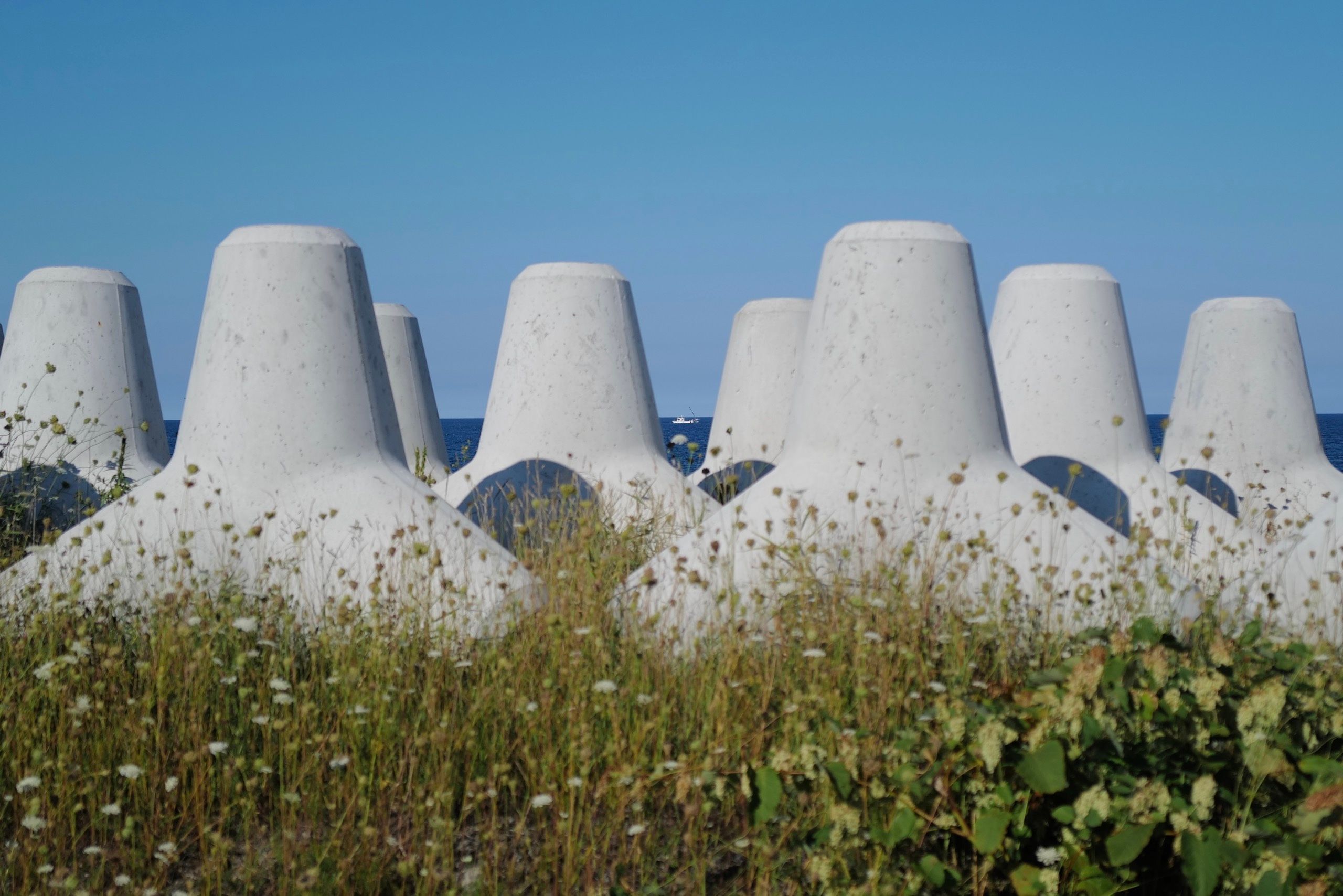 A tiny white fishing boat is visible through a line of concrete tetrapods on a beach