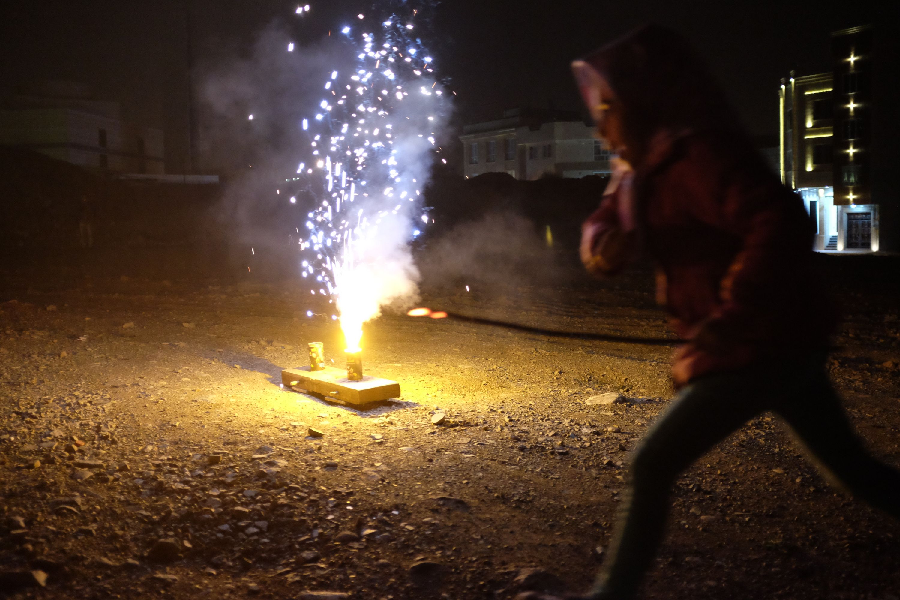 A small girl runs past a large sparkler placed on the ground, on a street at night.