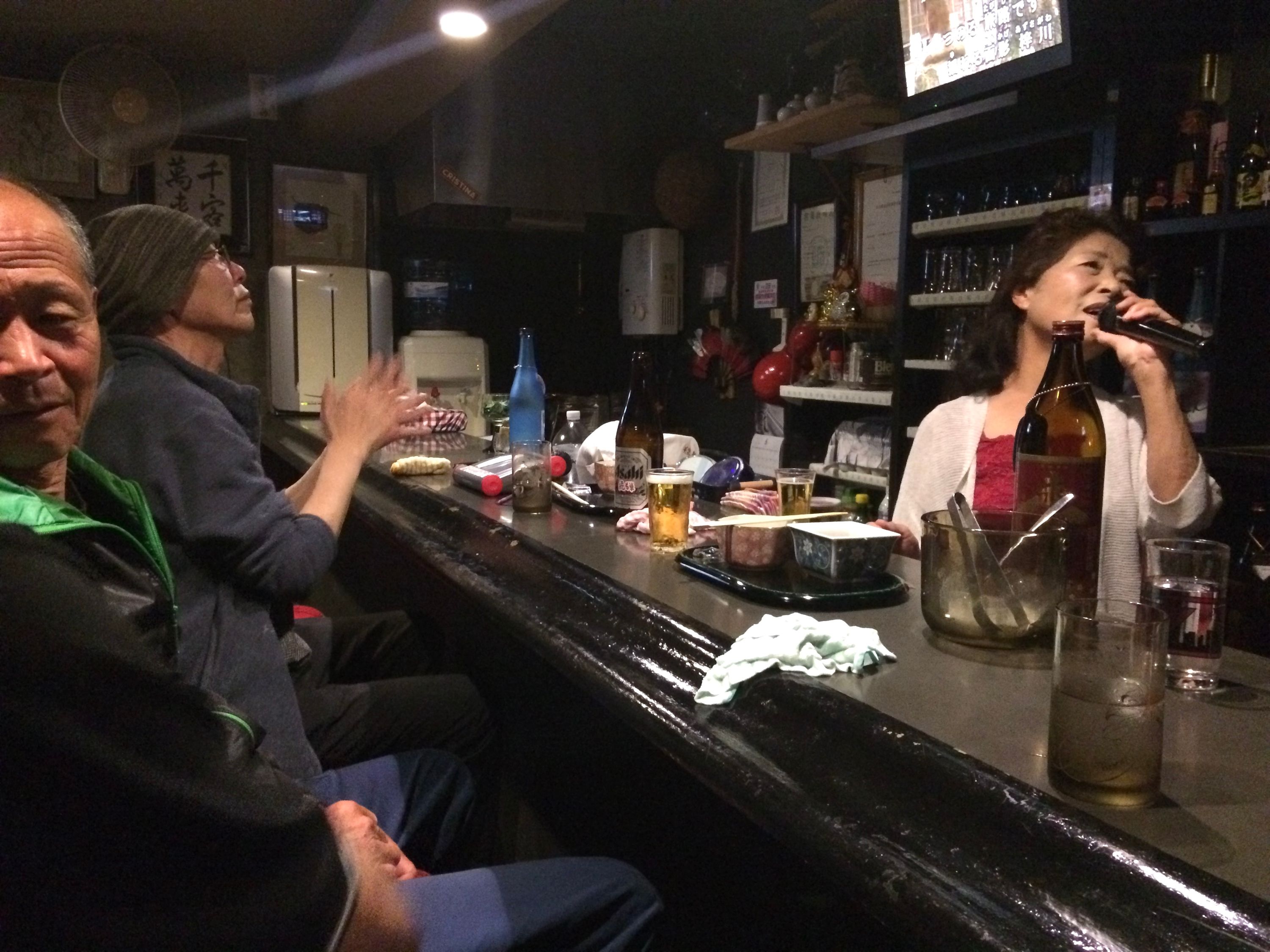 A female bartender sings in a karaoke bar, with male customers looking on.