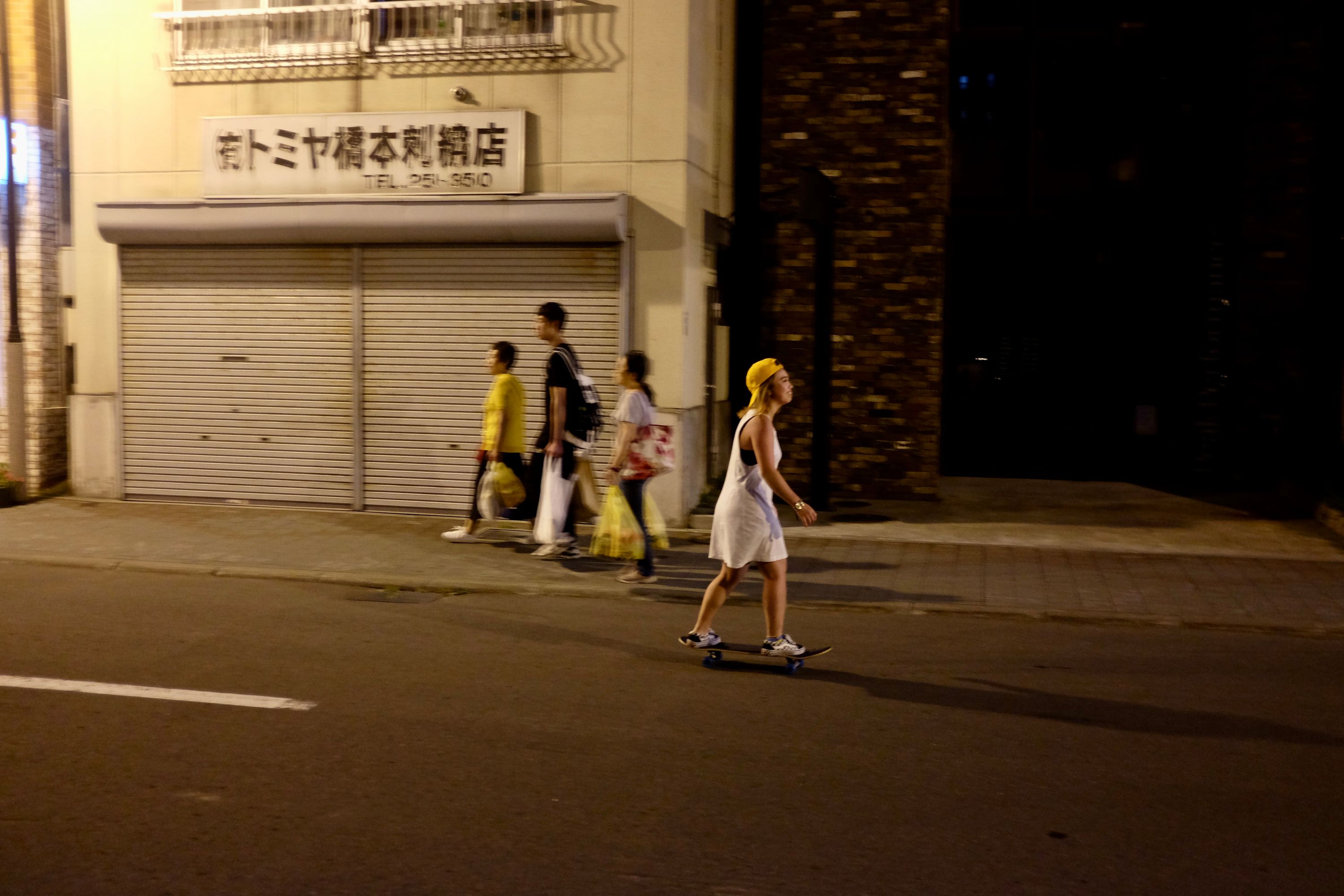 A Japanese girl in a yellow cap rides a skateboard down a street.