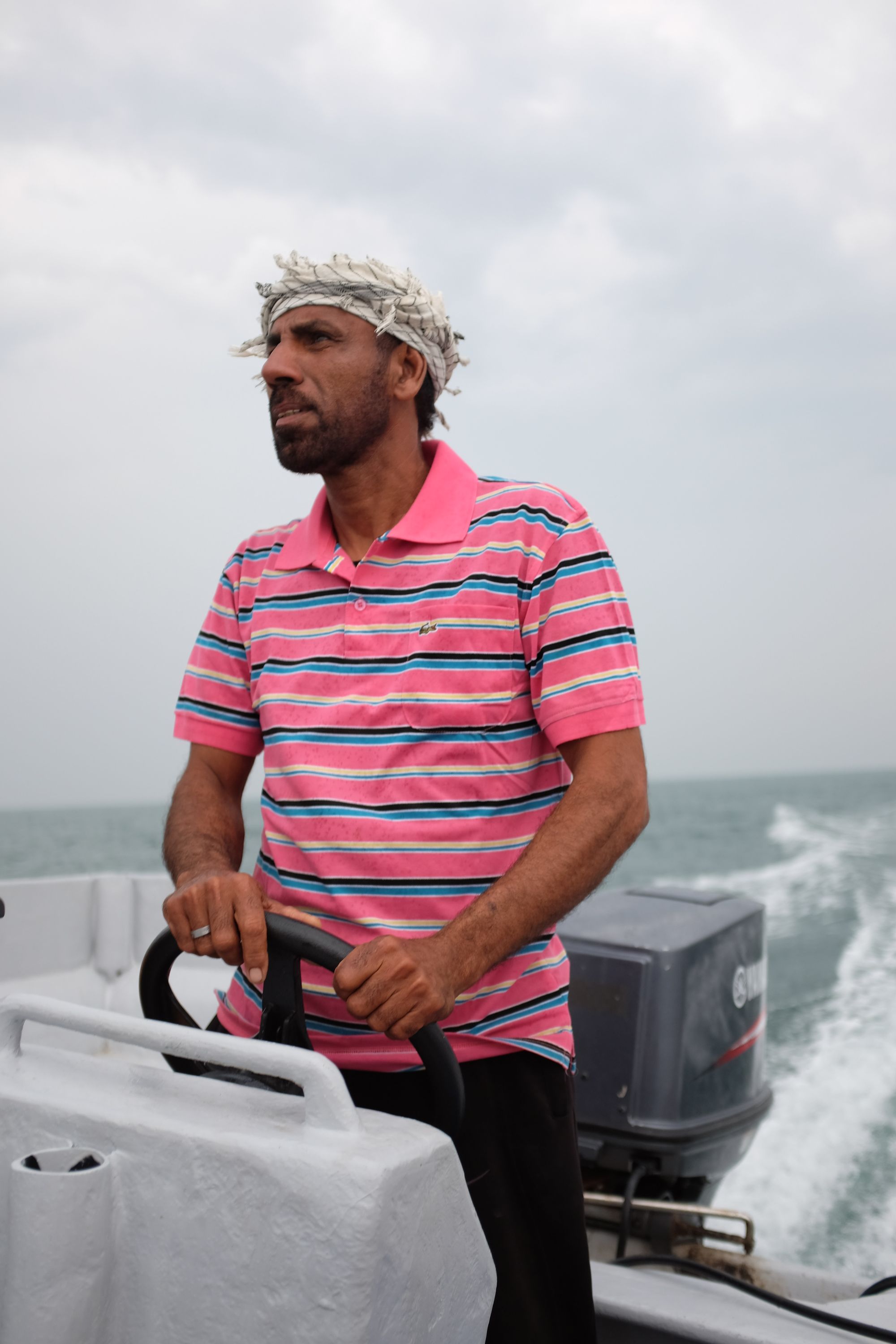 A man in a pink striped polo shirt with a scarf wrapped around his head drives a powerboat under a cloudy sky.