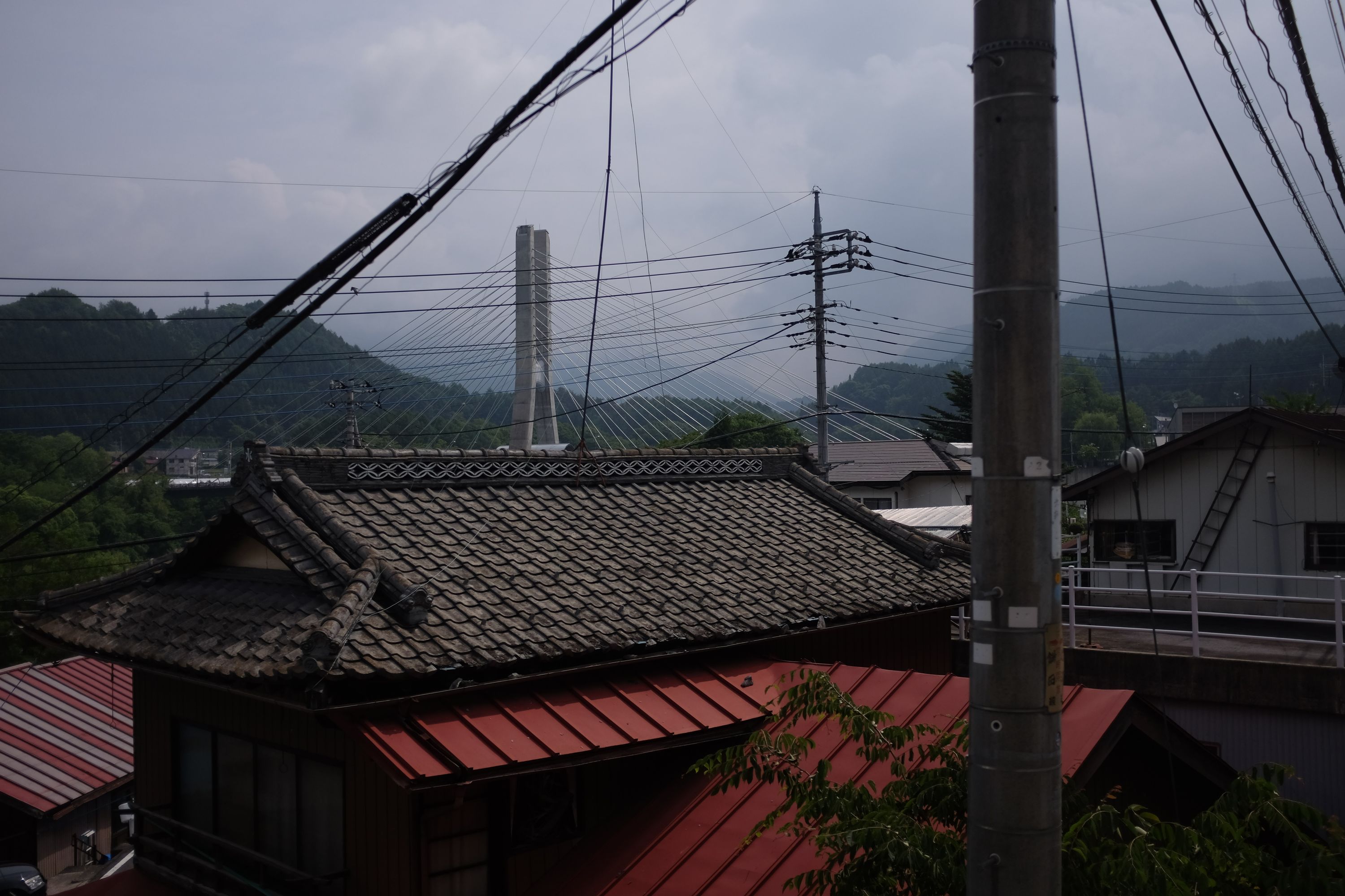 Looking towards the mountains over the roof of a Japanese house, lots of cables, and a small suspension bridge.
