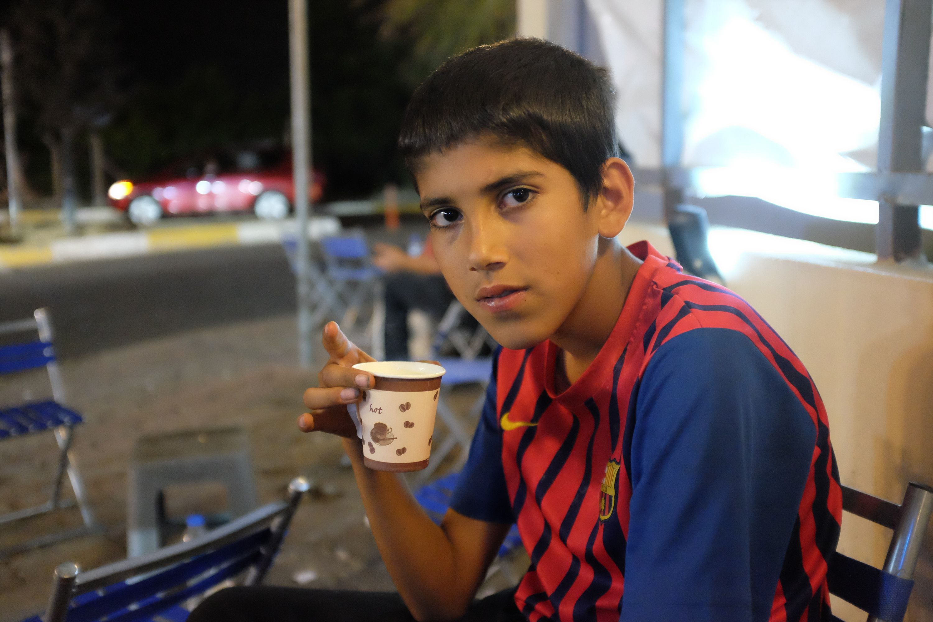 A young boy in an FC Barcelona shirt holds a paper cup of tea, sitting on a chair by the side of a road.